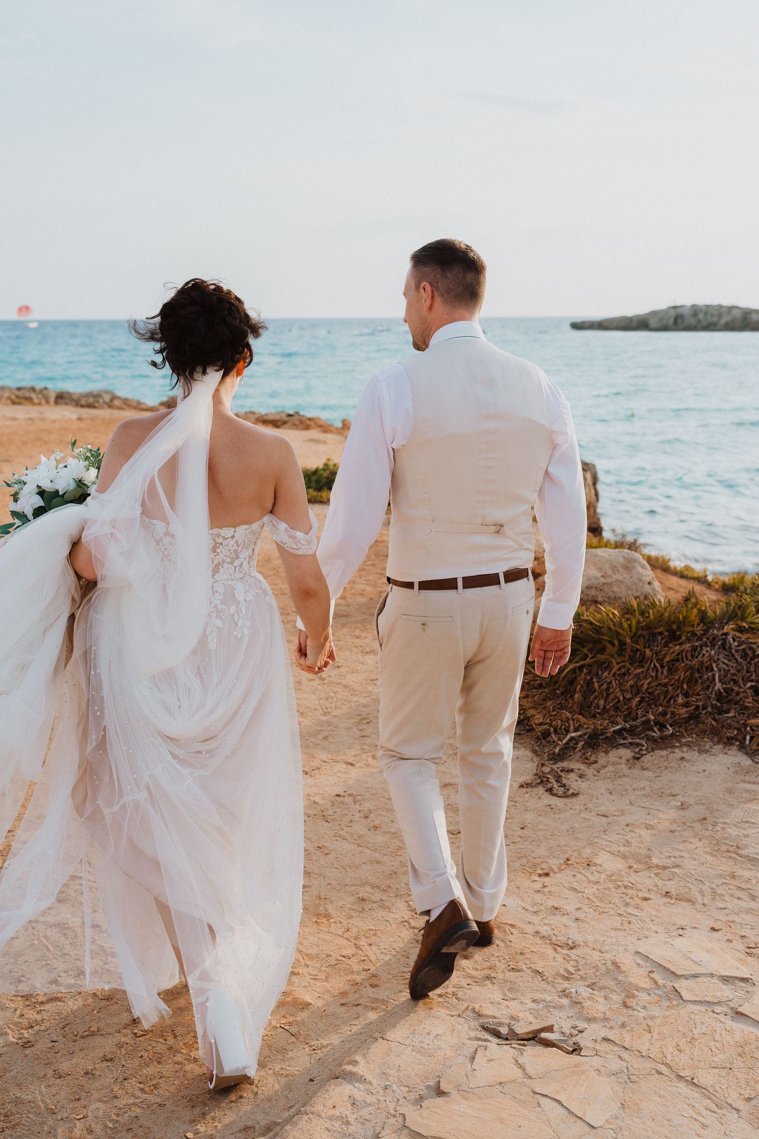 A couple in wedding attire walks hand in hand along a rocky coastal shoreline during a sunset beach wedding.