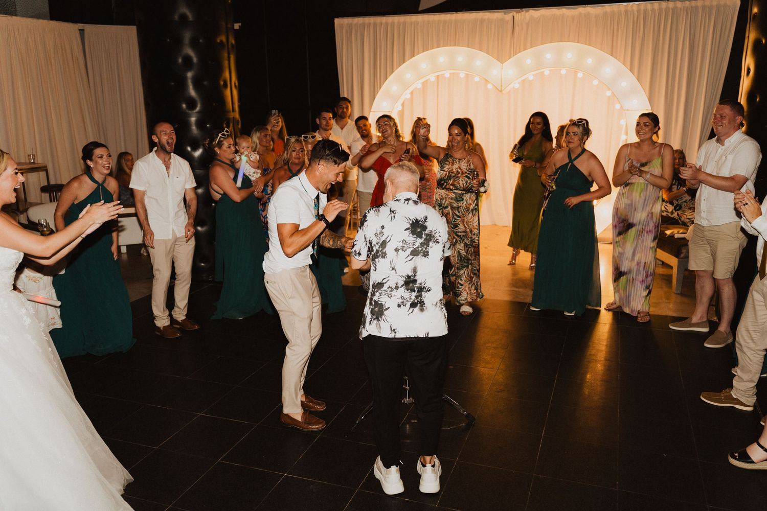 Wedding guests dancing together on a dark dance floor at a reception with people in green dresses and white attire.