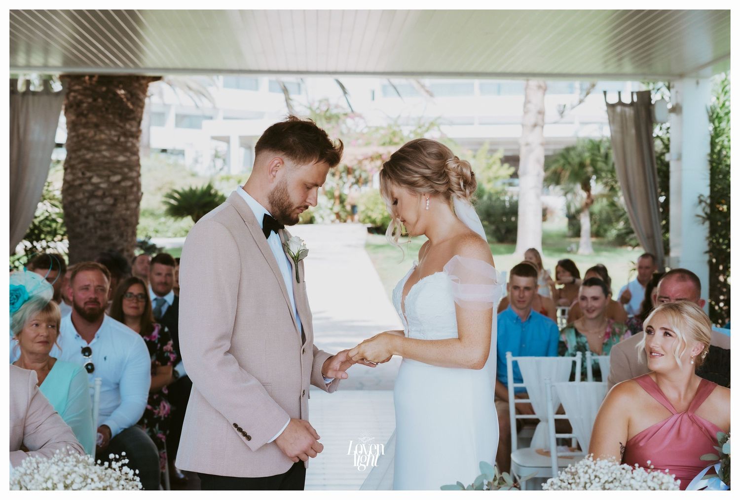 Intimate moment during ring exchange at an outdoor wedding ceremony with guests looking on.