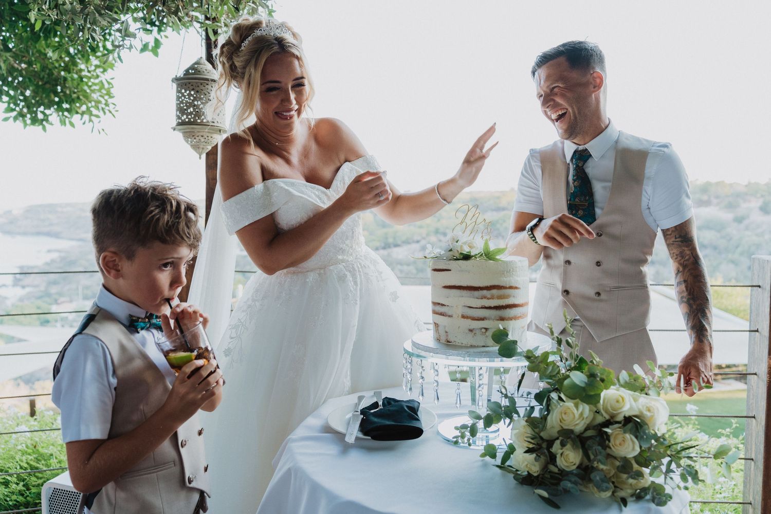 Wedding party gathers around cake table at outdoor venue with greenery and flowers while enjoying refreshments.