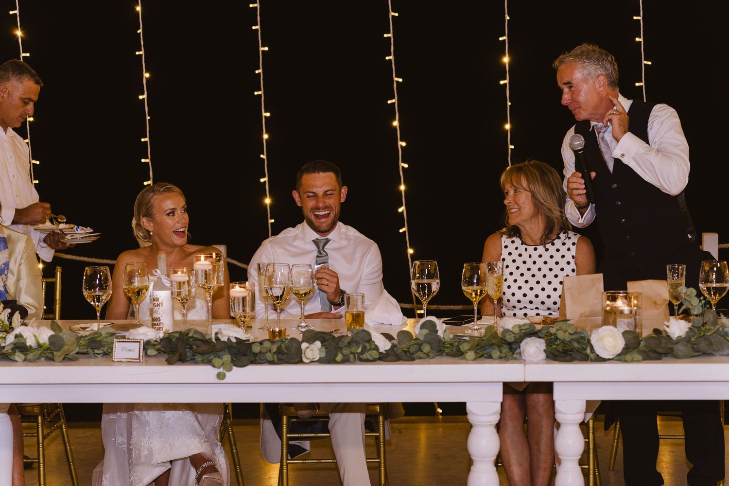 Wedding party enjoys speeches and toasts at elegant head table decorated with greenery.
