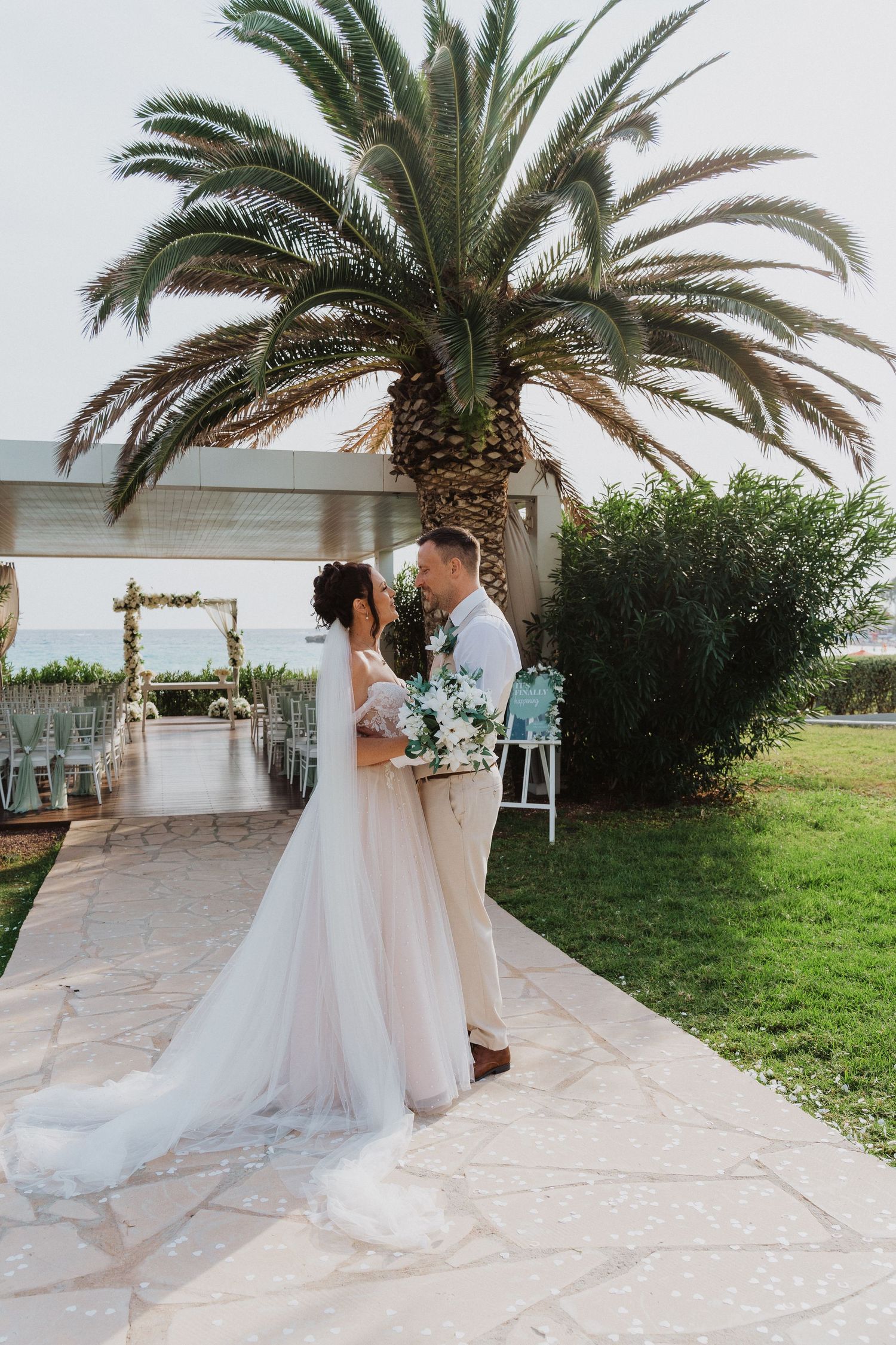A couple embraces at an outdoor wedding ceremony with a palm tree and decorated arch against a coastal backdrop.