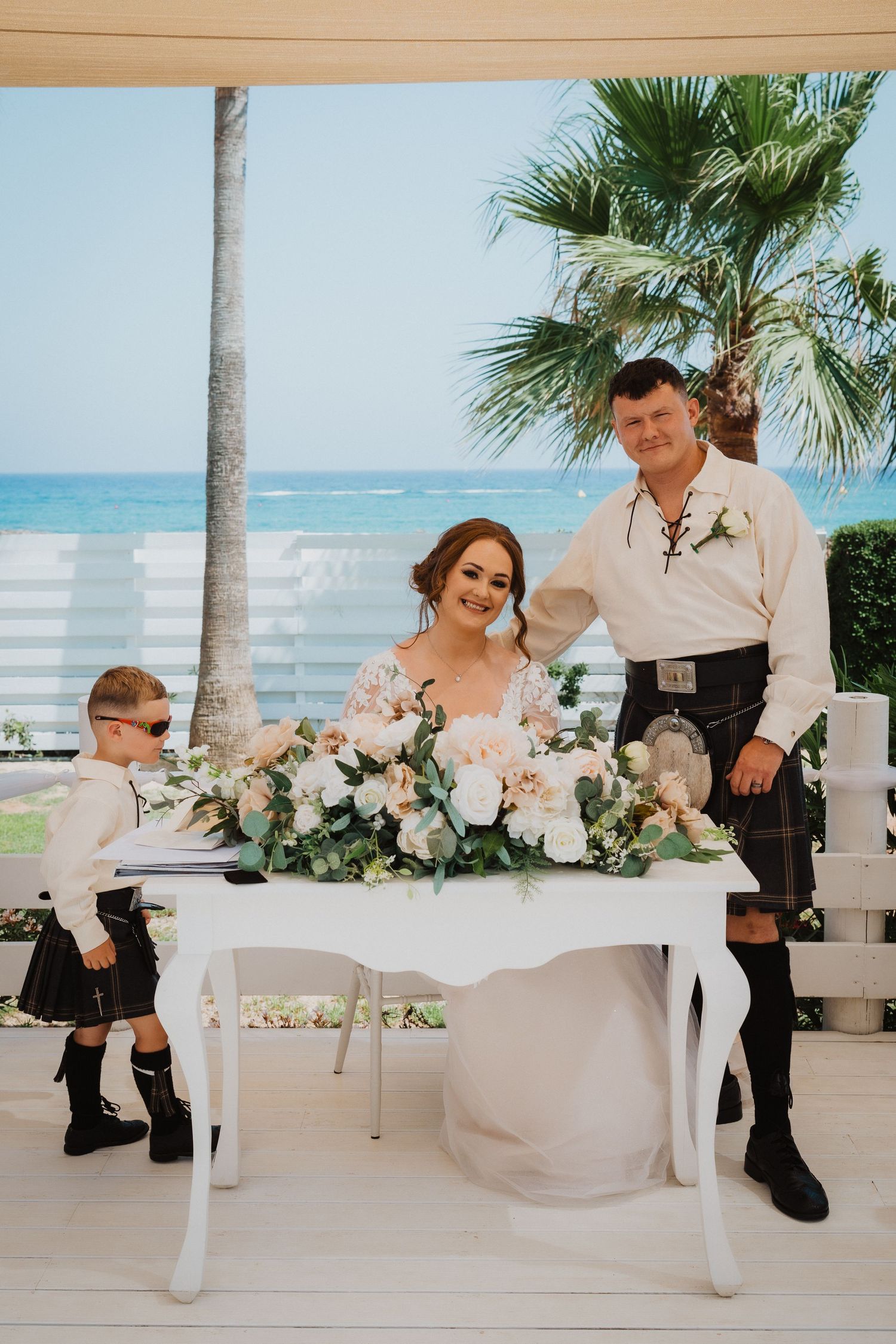 Scottish-themed beach wedding with groom in kilt, bride in lace dress, and young attendant at signing table.