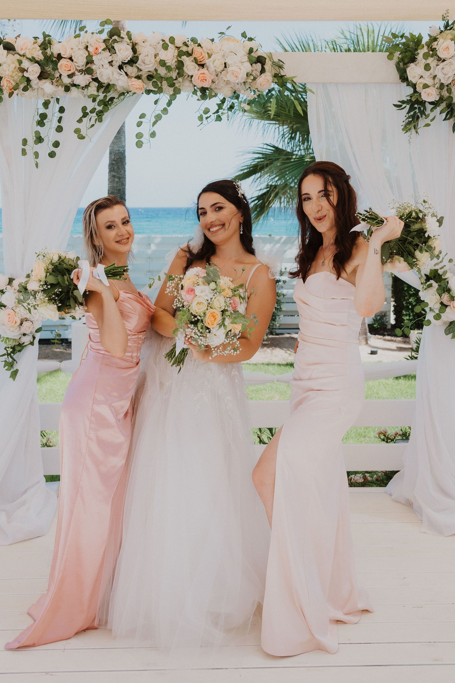 Three women in flowing pink and white dresses pose under floral wedding arch by the beach.