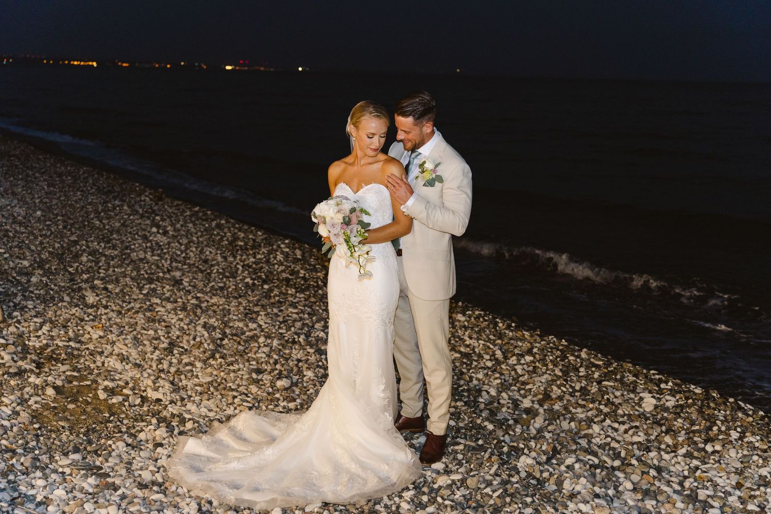 Romantic nighttime beach wedding portrait with couple embracing on rocky shoreline.