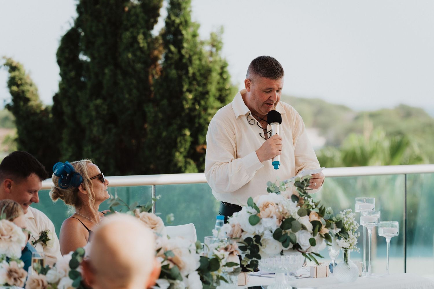 A speaker delivers a toast during an elegant outdoor wedding reception with white floral arrangements.
