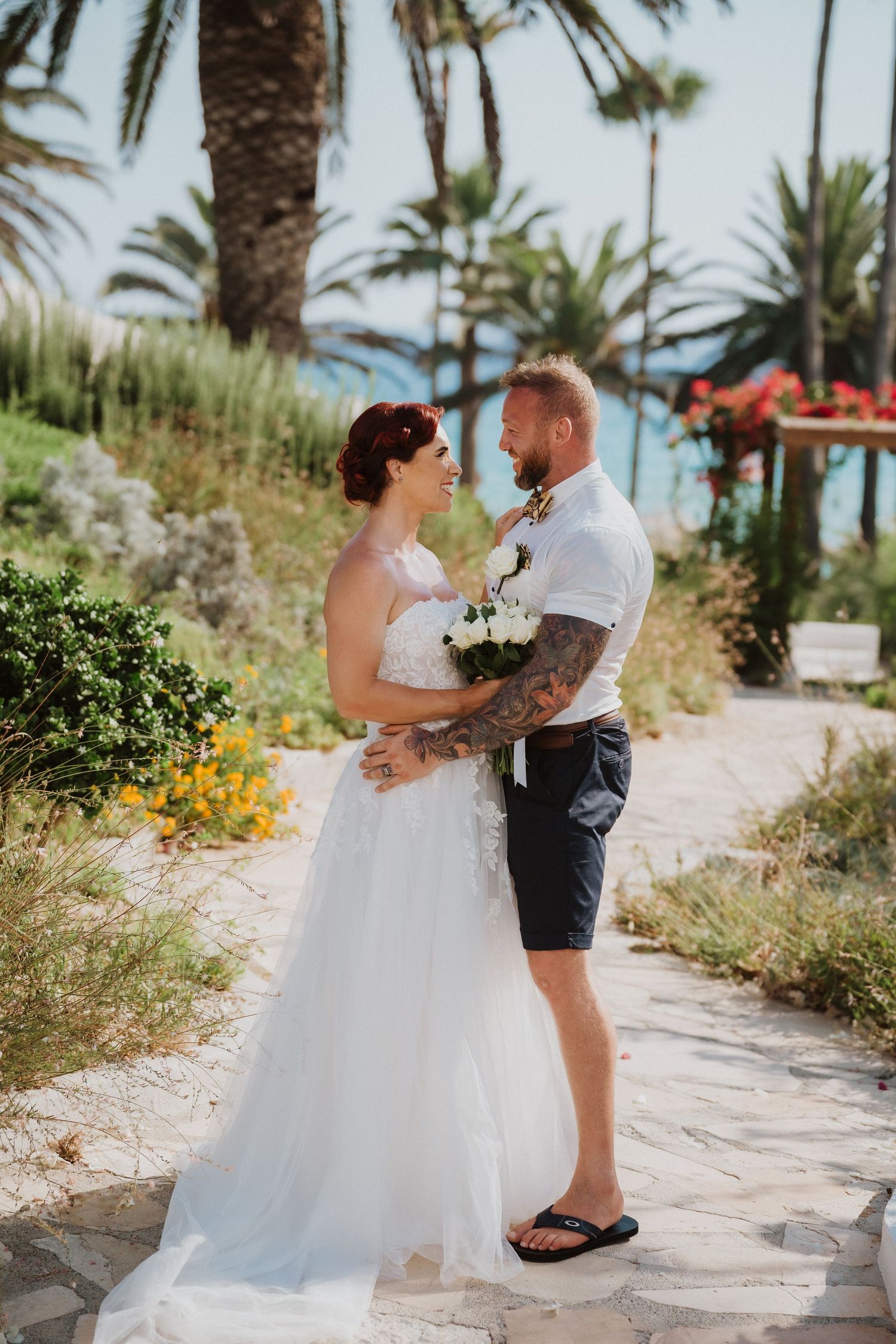 A couple embraces on a tropical garden path with palm trees and ocean views in the background.