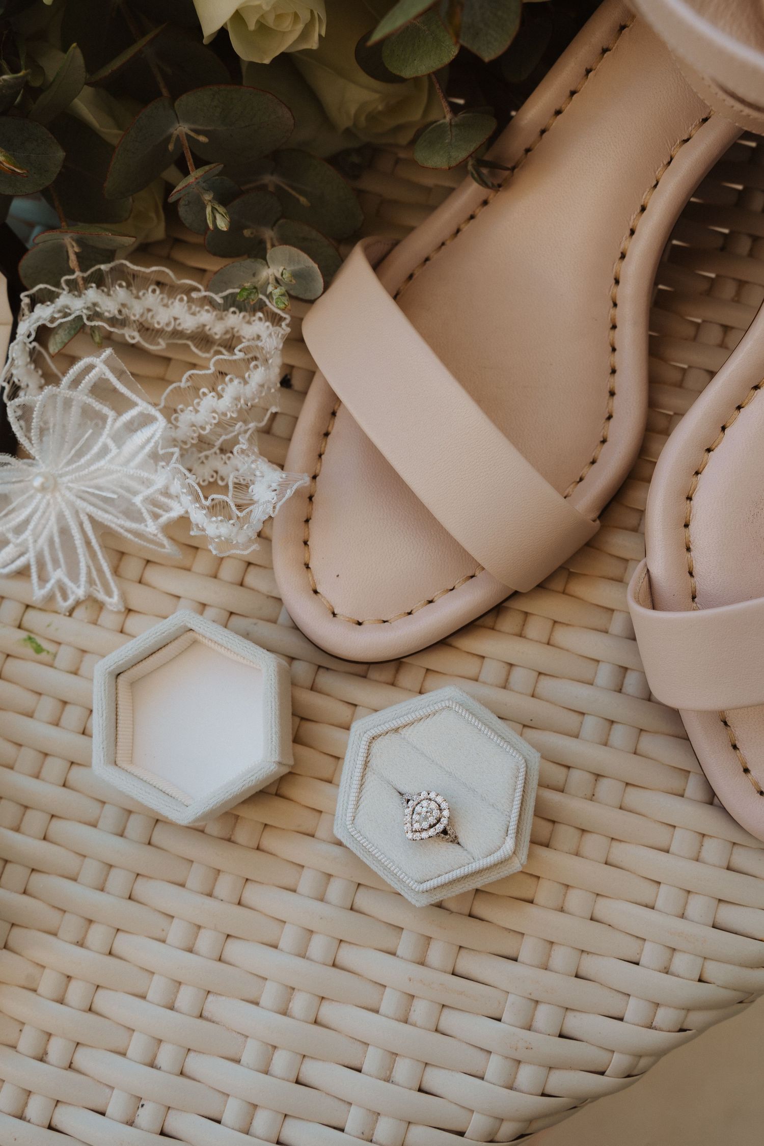 Wedding accessories including beige sandals, lace details, and ring boxes arranged on a woven surface.