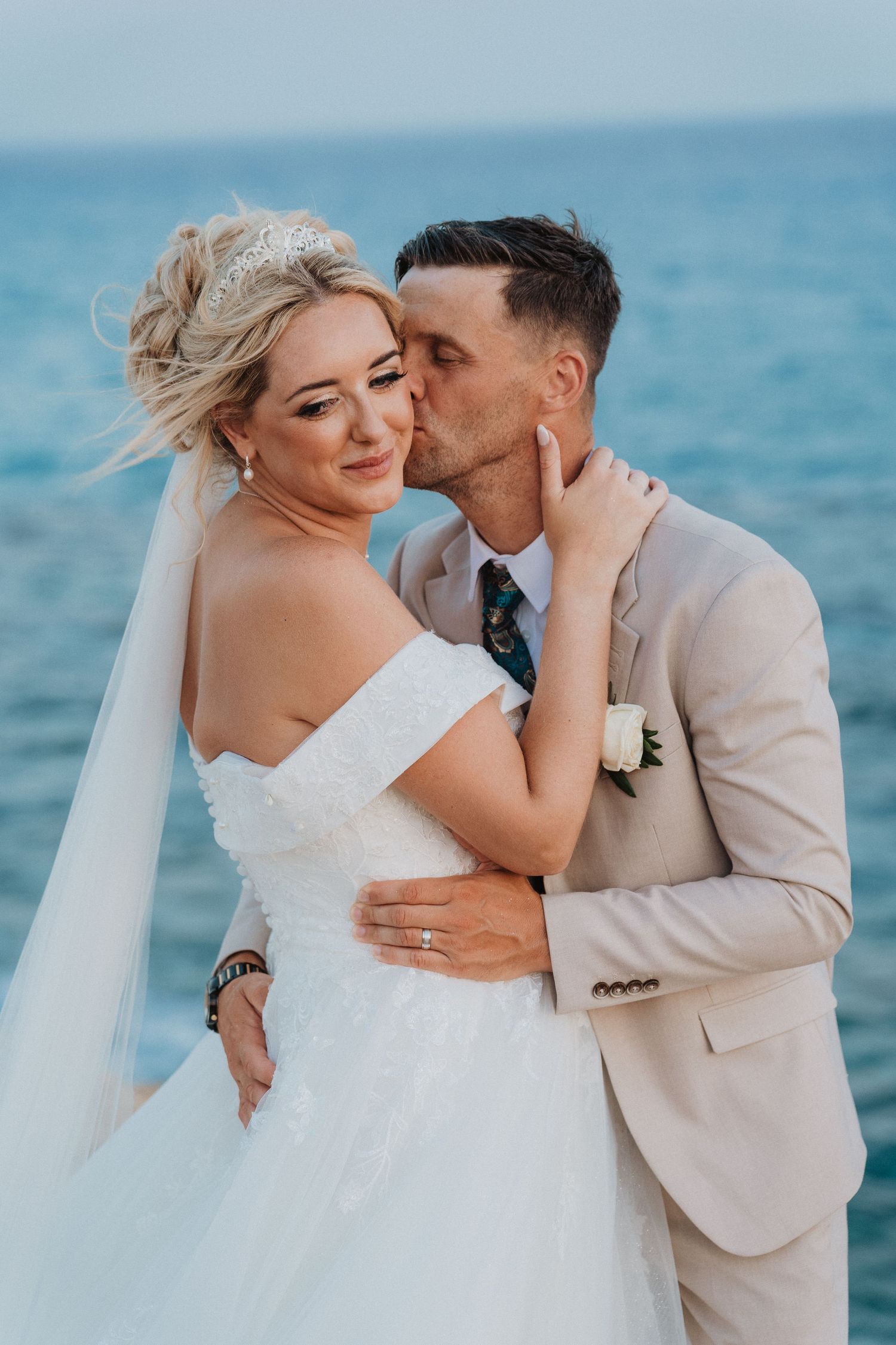 A romantic wedding couple embraces by the ocean, with the bride in white and groom in beige.