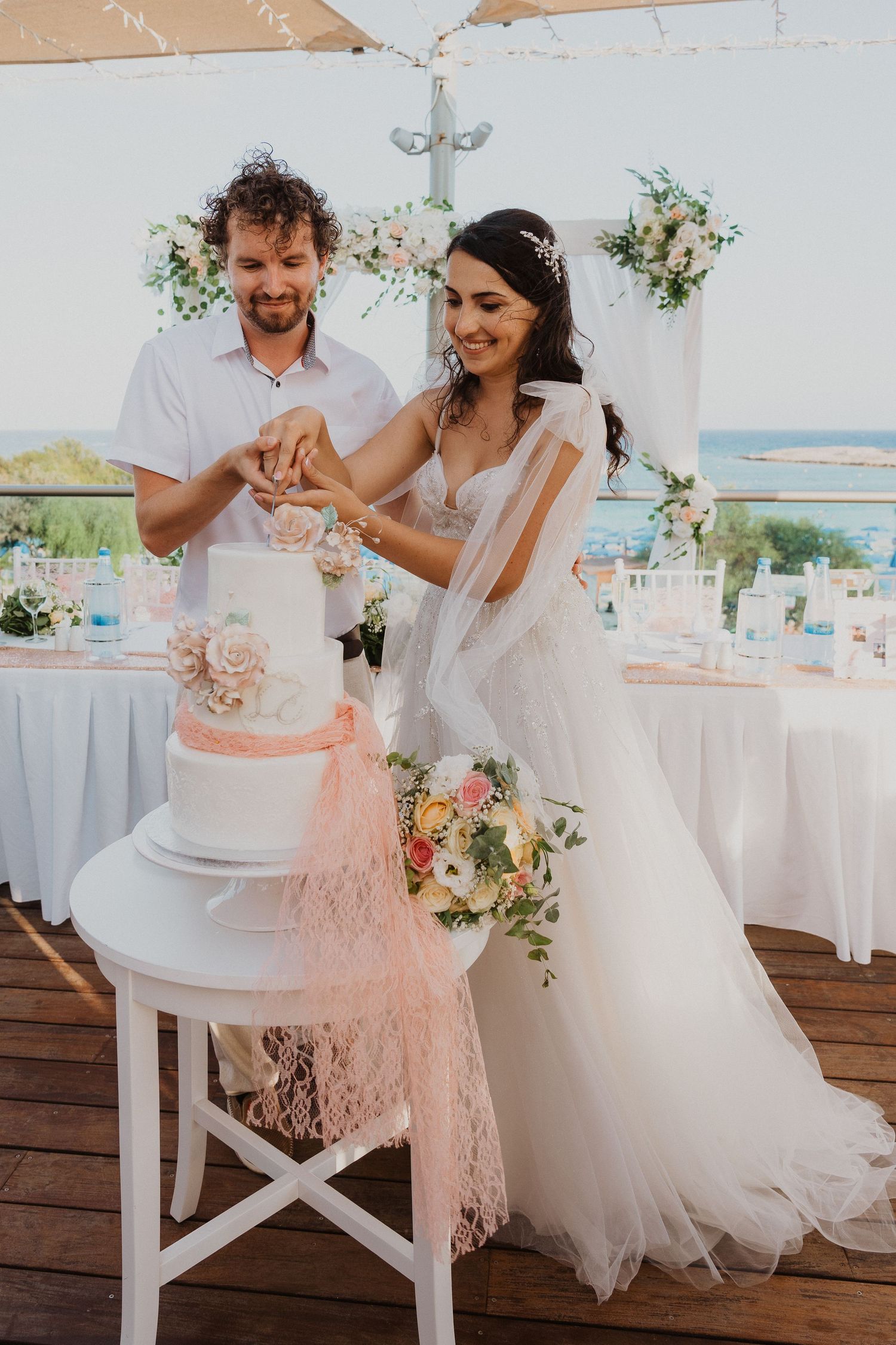 A couple cuts their elegant white and pink wedding cake together at an outdoor beach reception.