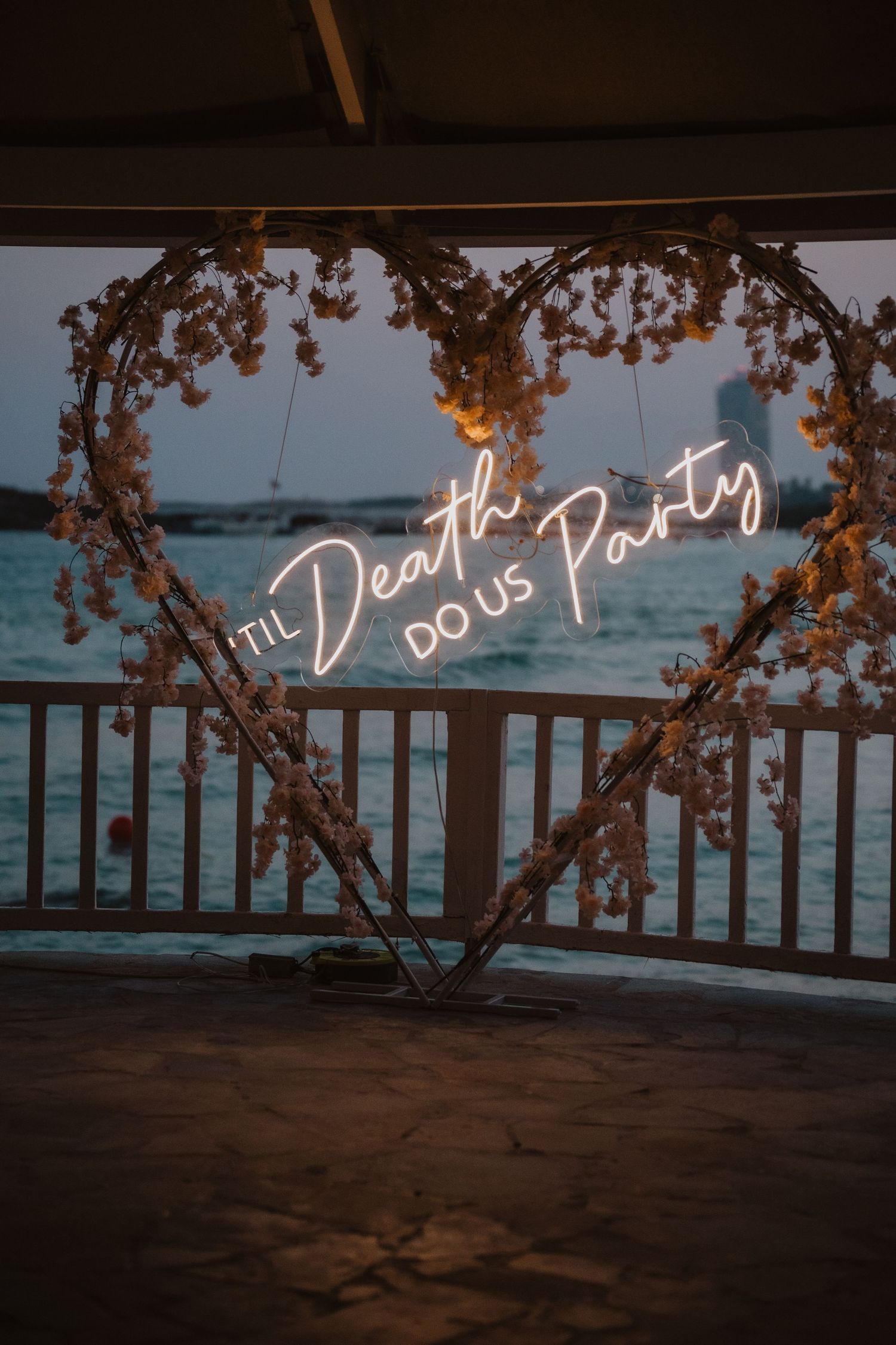 Heart-shaped floral arch with neon 'Til Death Do Us Party' sign overlooking ocean at dusk.