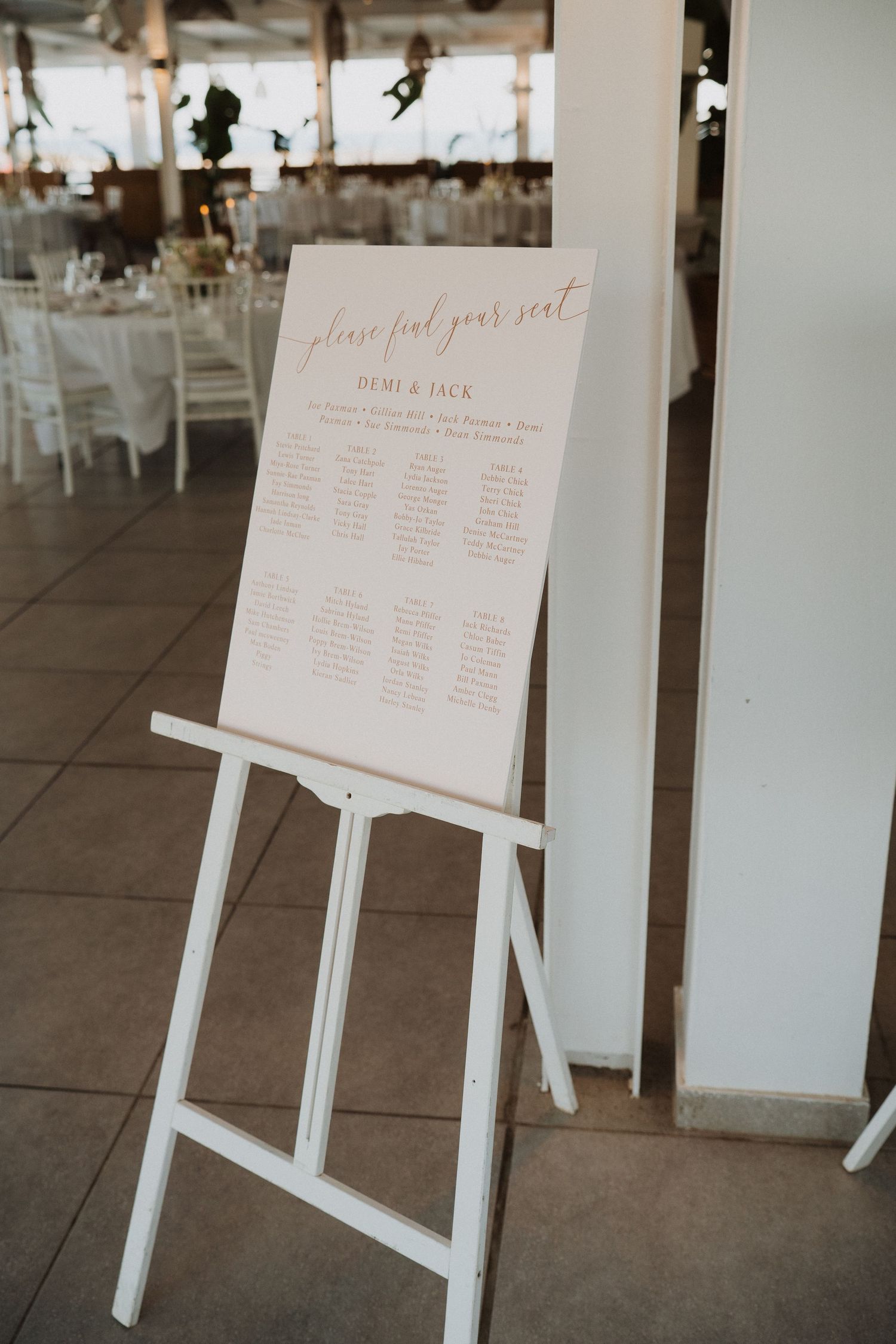 Wedding reception seating chart signs displayed on white easels in an elegant venue with tiled floors.