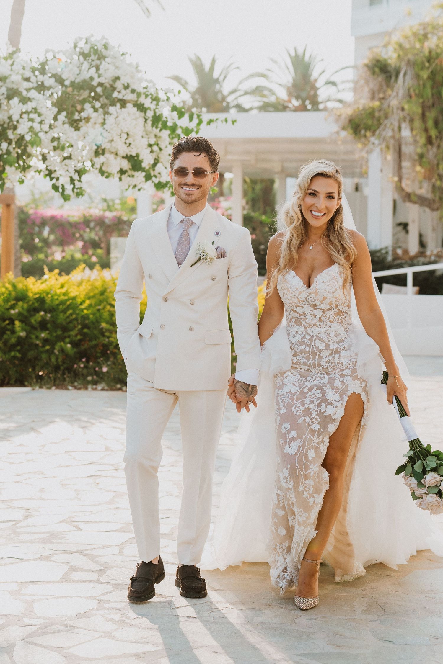 A couple in elegant white wedding attire pose together on a stone pathway with tropical foliage in the background.