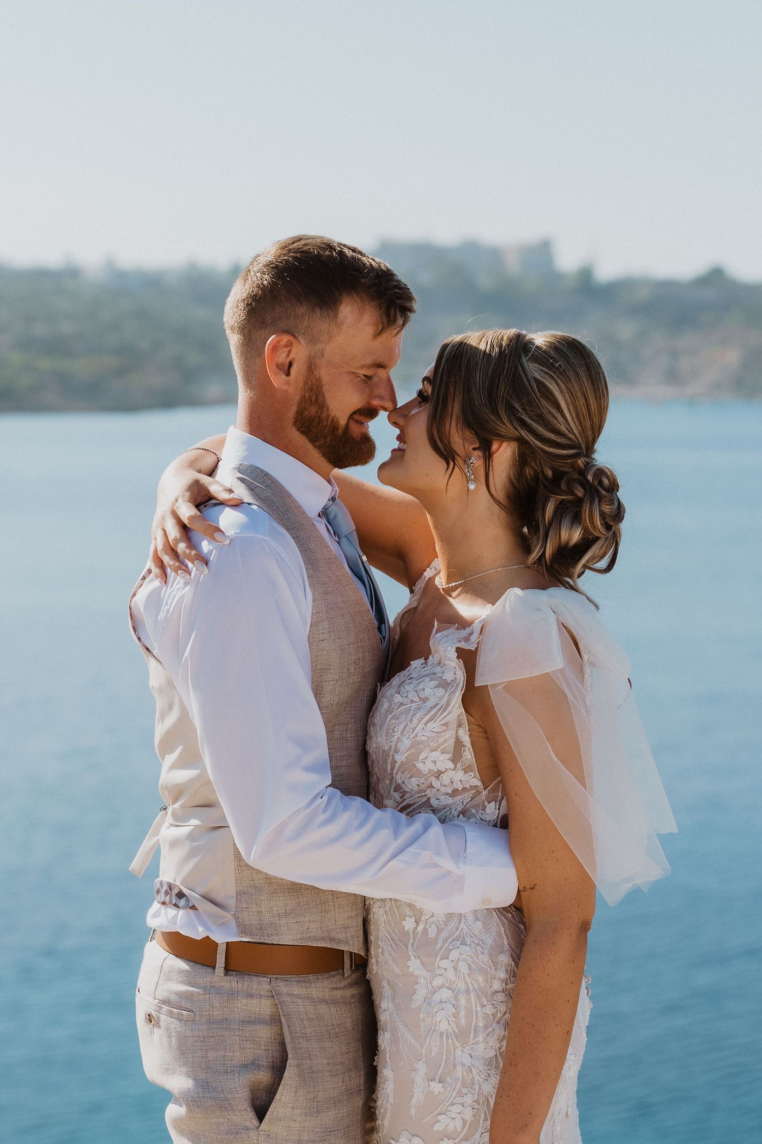 Newlyweds share intimate moment by seaside in elegant wedding attire.
