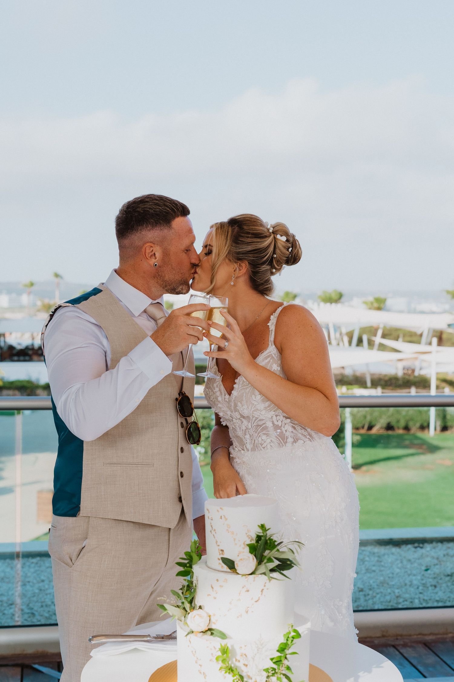 A couple shares a romantic kiss during their wedding celebration on a waterfront deck with white umbrellas in the background.