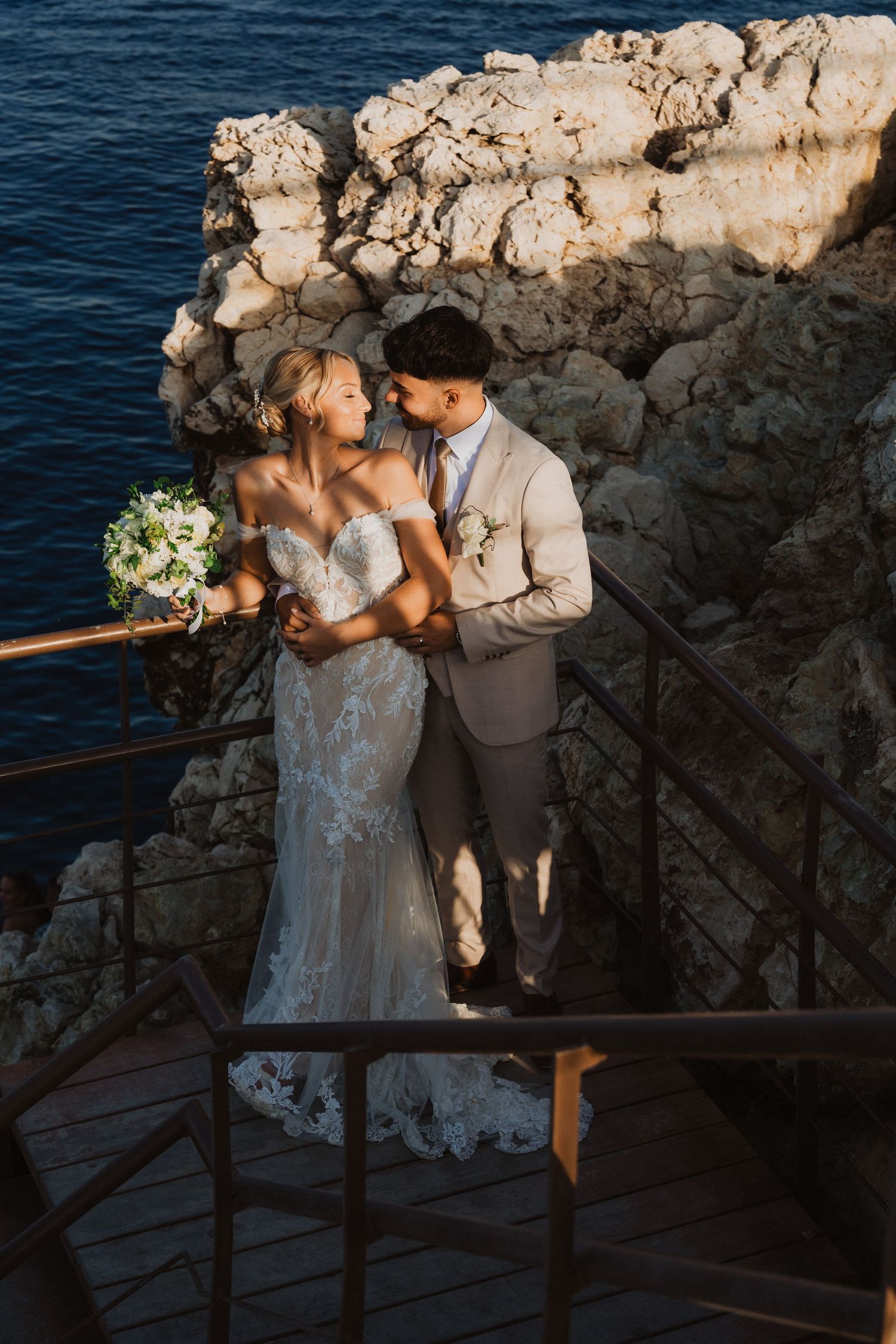 Intimate wedding portrait on seaside deck with rocky cliffs and ocean backdrop.