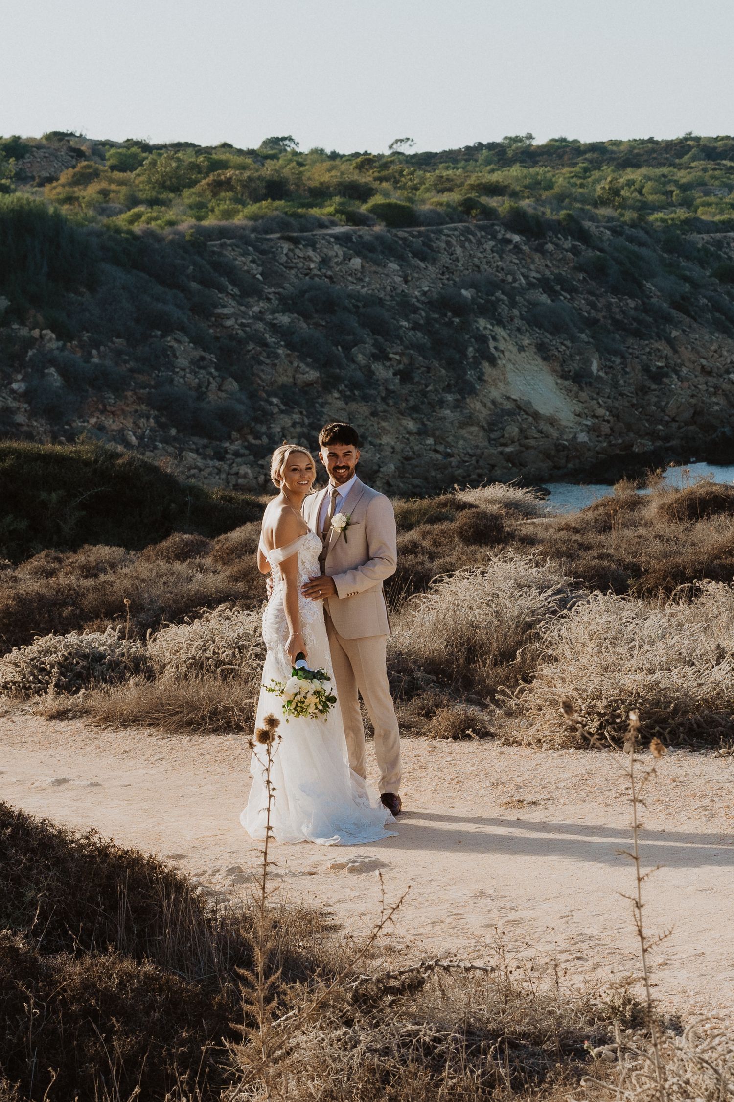 Couple walking together on a sunlit coastal path with dried brush and ocean views.