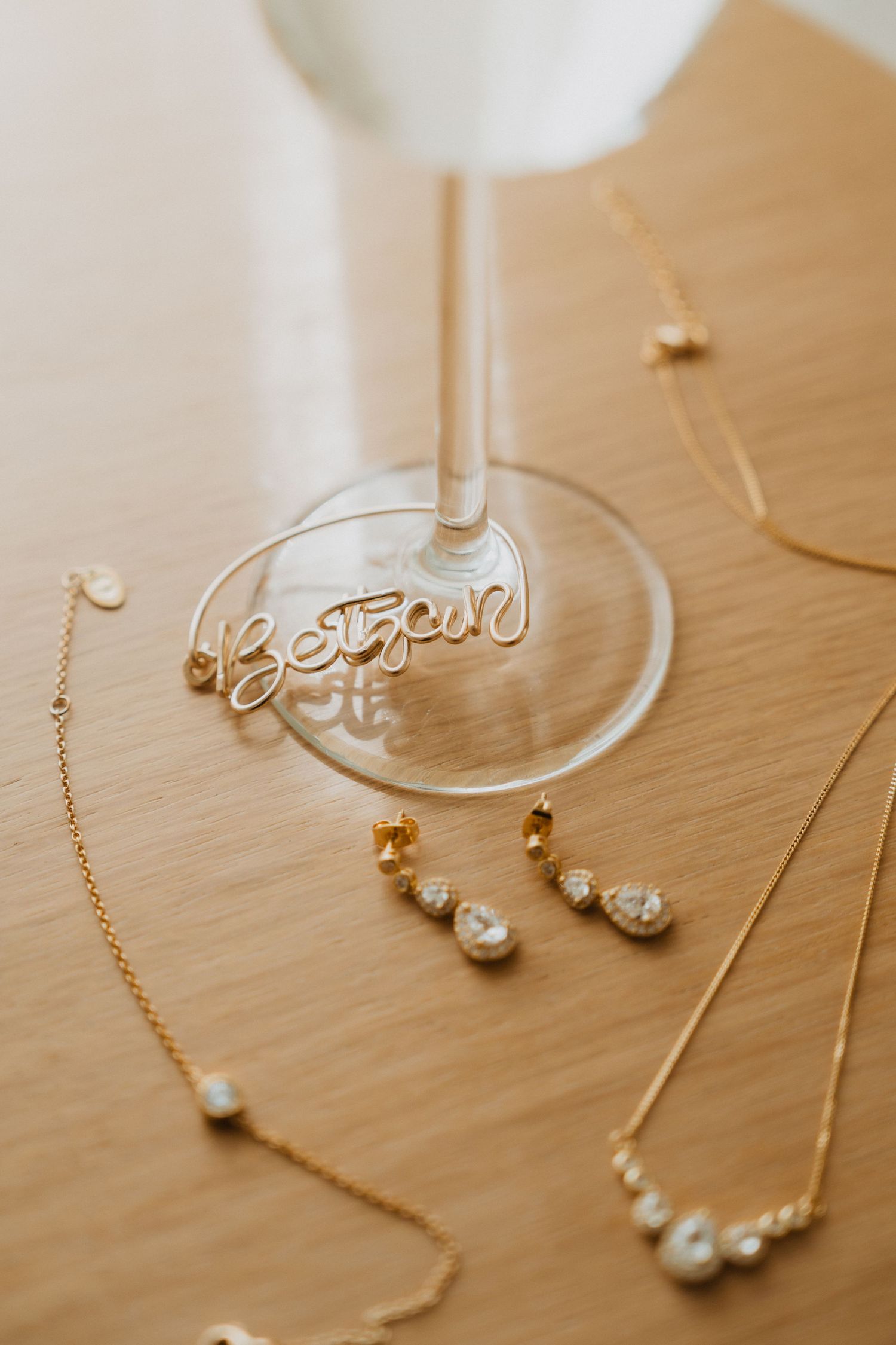 Wedding jewelry and accessories arranged around a personalized champagne glass on a wooden surface.