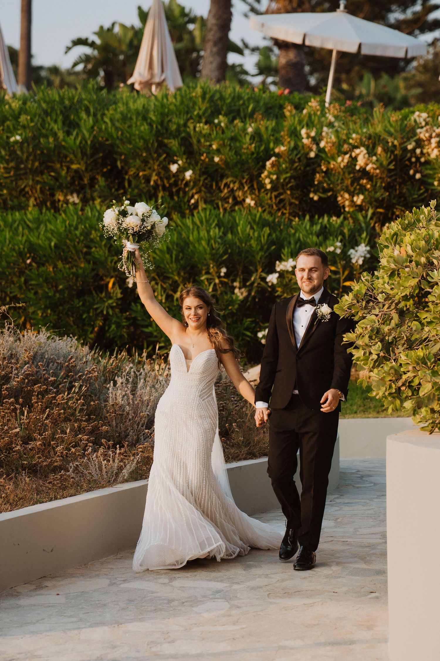 Joyful bride raises bouquet while walking hand-in-hand with groom through lush garden at golden hour.