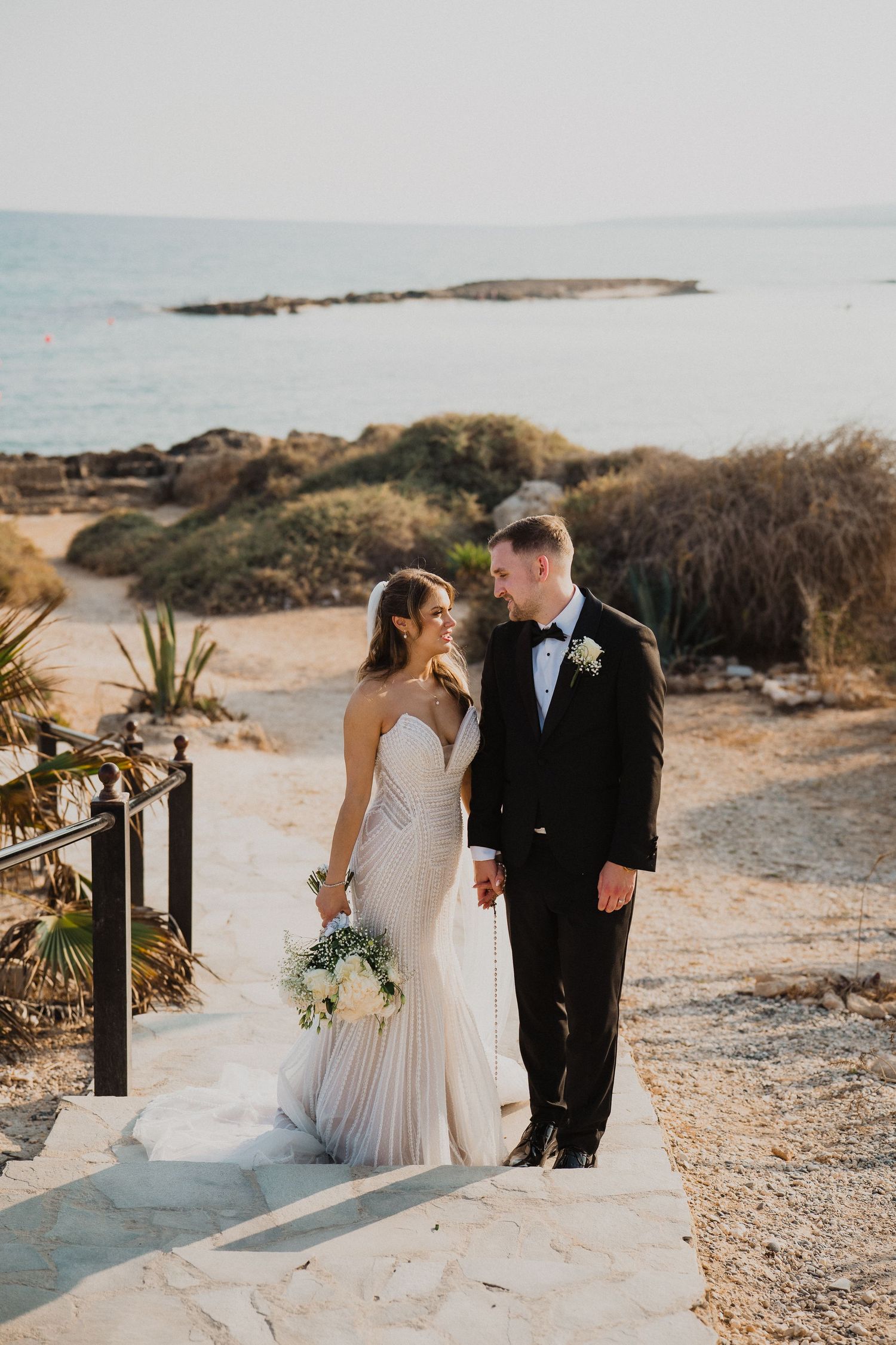 Newlyweds walking hand in hand along a coastal stone path with the blue sea in the background.