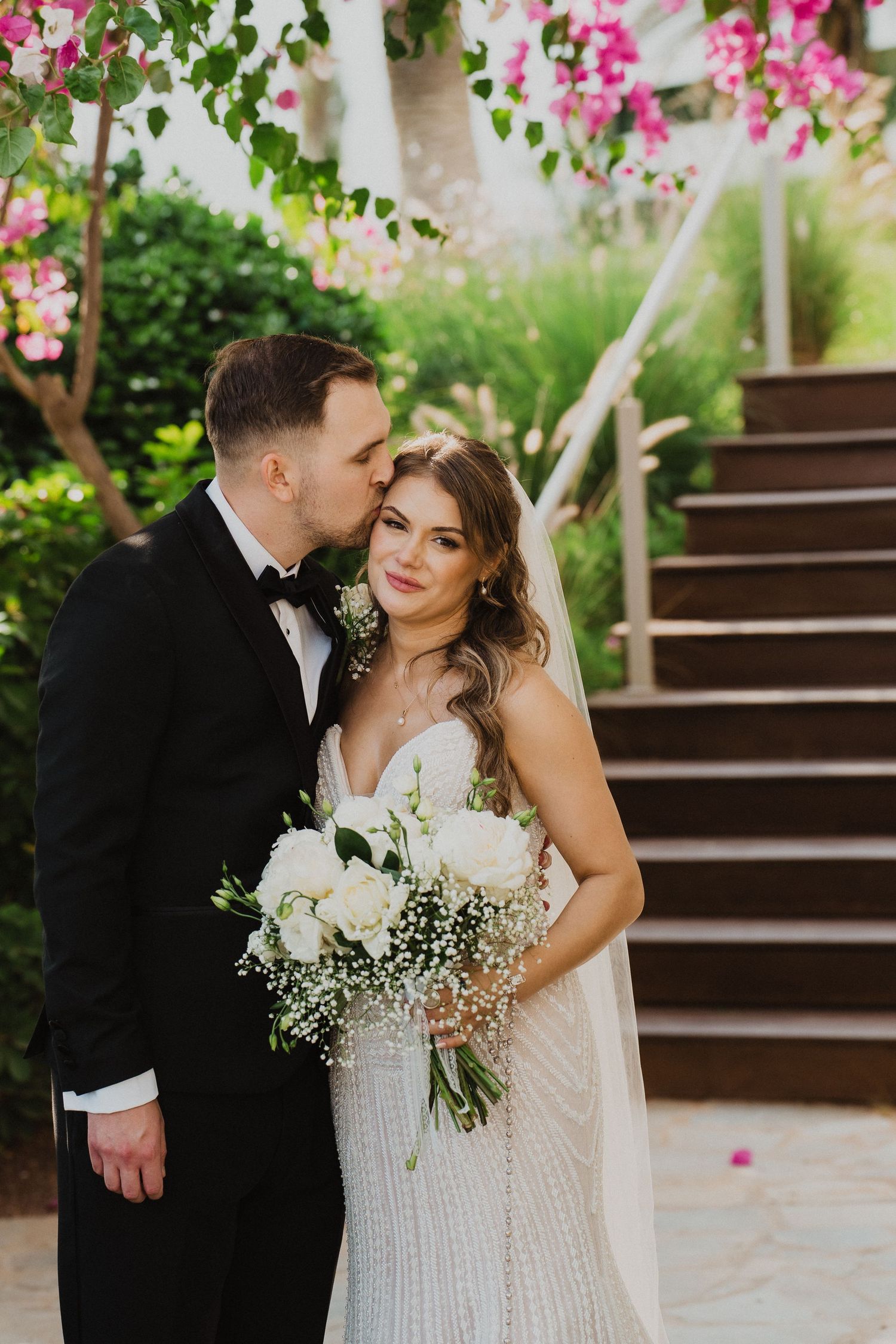 Groom kisses bride on cheek as she smiles holding white rose bouquet under pink bougainvillea.