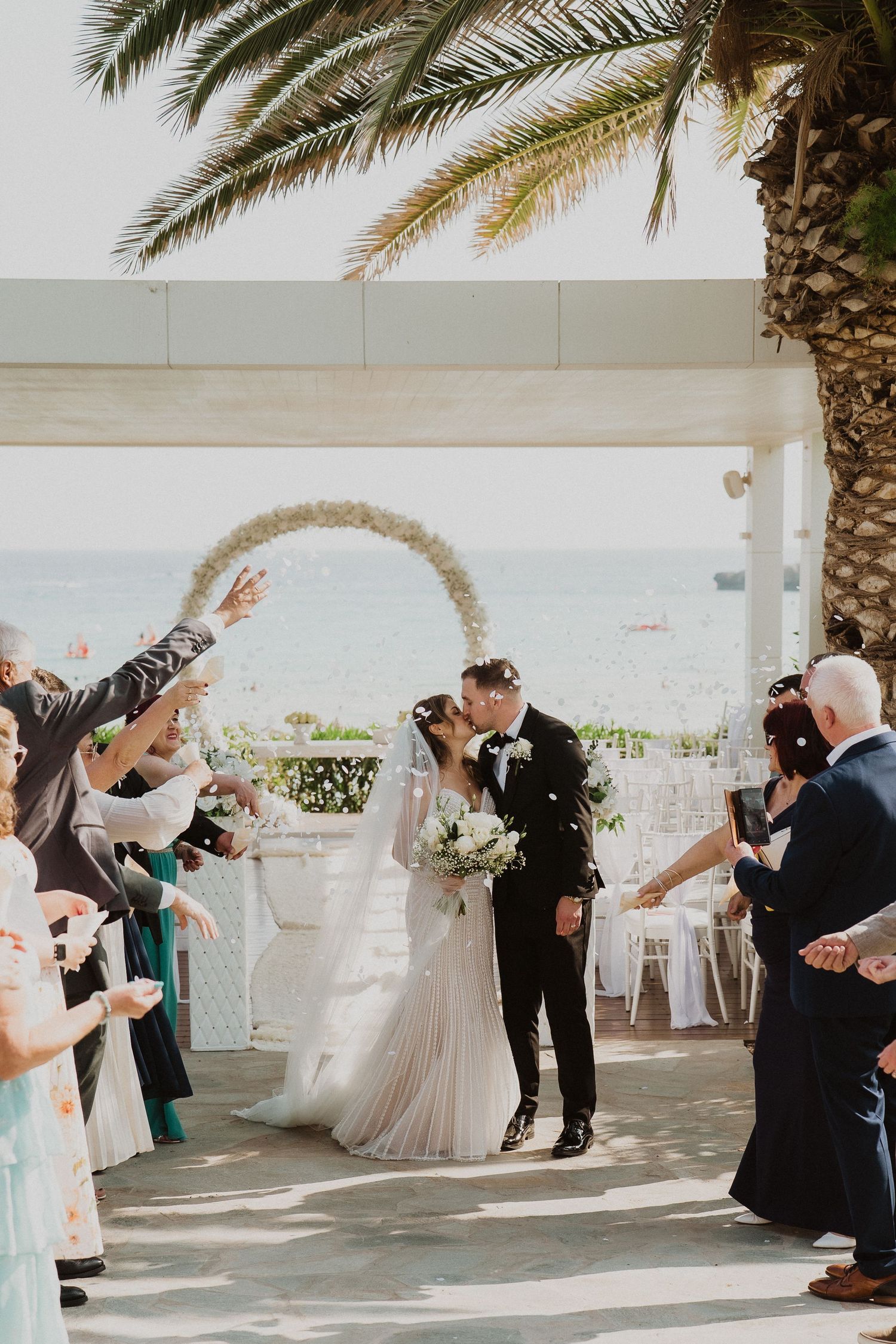 Bride and groom share a kiss during their outdoor wedding ceremony by the sea, surrounded by cheering guests under a floral arch.
