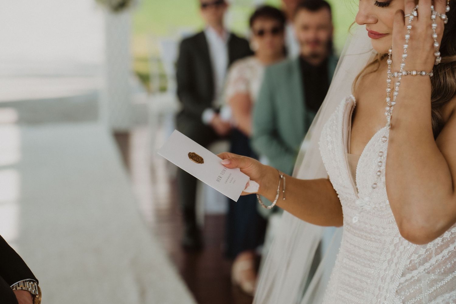 Bride reading vows from paper during outdoor wedding ceremony with guests seated behind.
