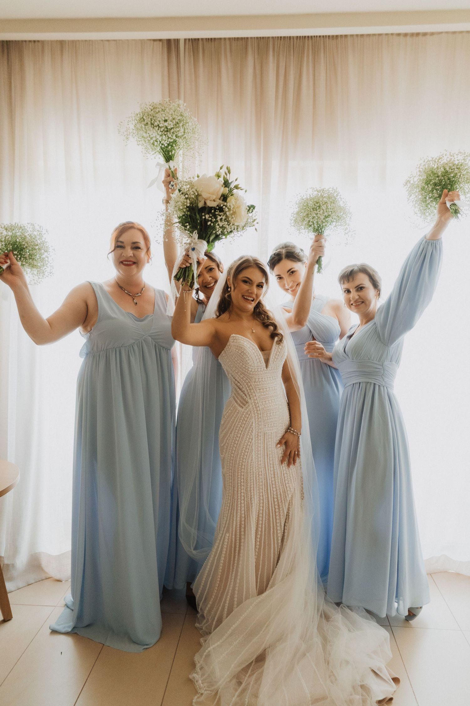 Bride in white gown with four bridesmaids in light blue dresses raising bouquets.