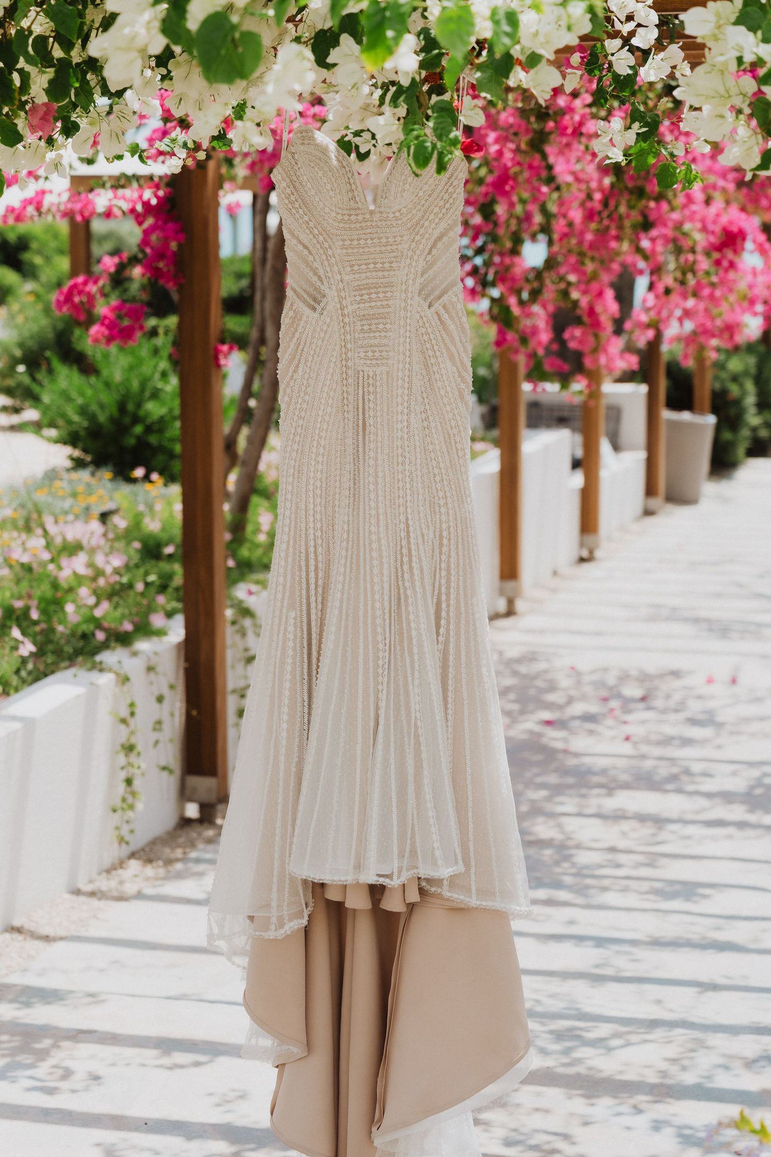 Elegant beige beaded wedding dress hanging outdoors among vibrant pink bougainvillea flowers.