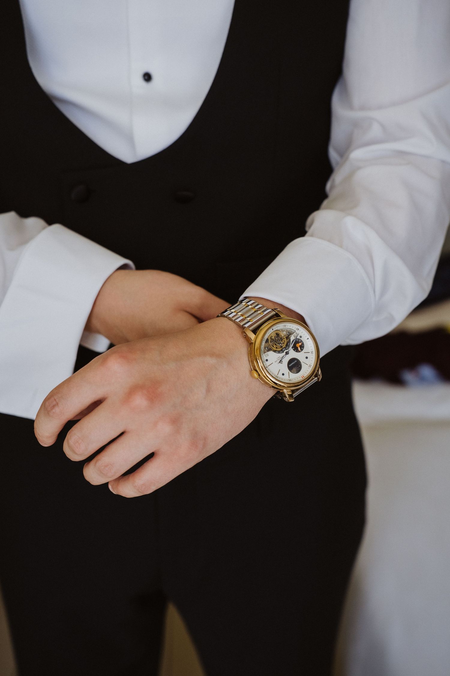 Groom adjusting his gold skeleton watch while dressed in a black tuxedo vest and white dress shirt.