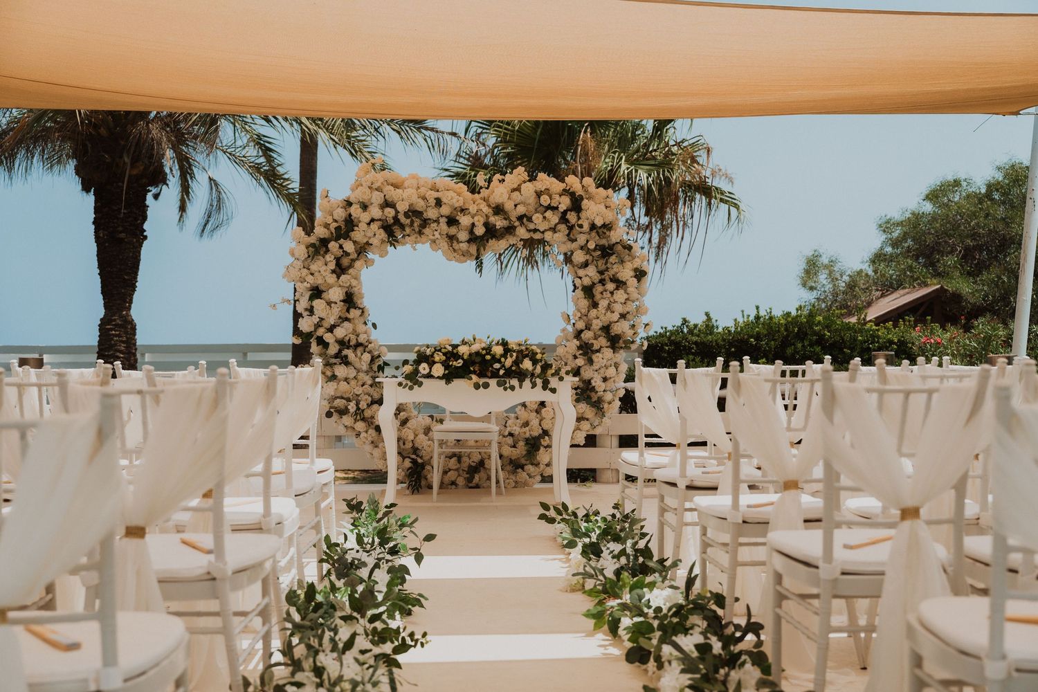 Elegant outdoor beach wedding ceremony with floral arch, white chairs, and tropical palm trees under a canopy.