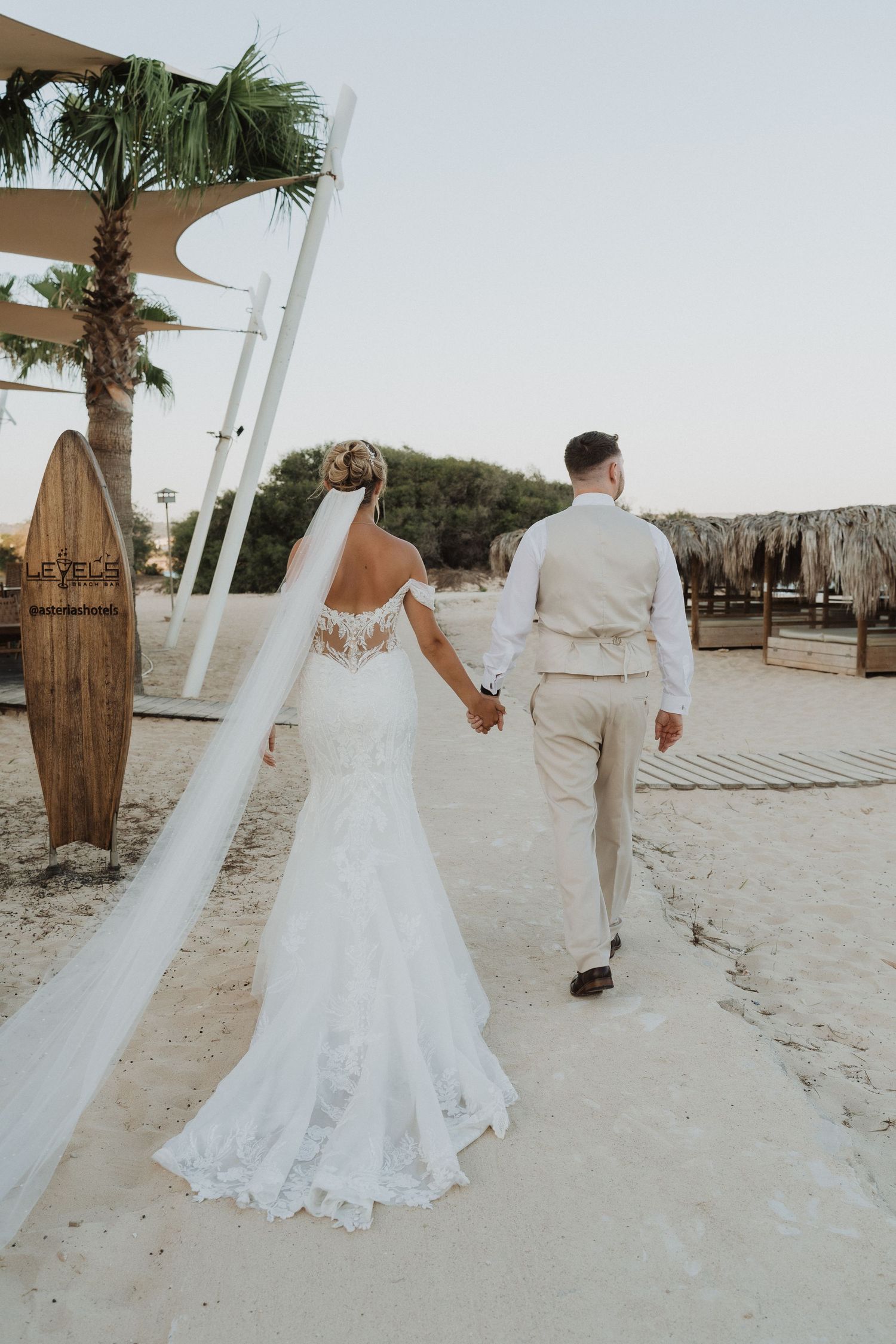 A couple walks hand in hand on a tropical beach during their wedding, with white decor and palm trees in the background.