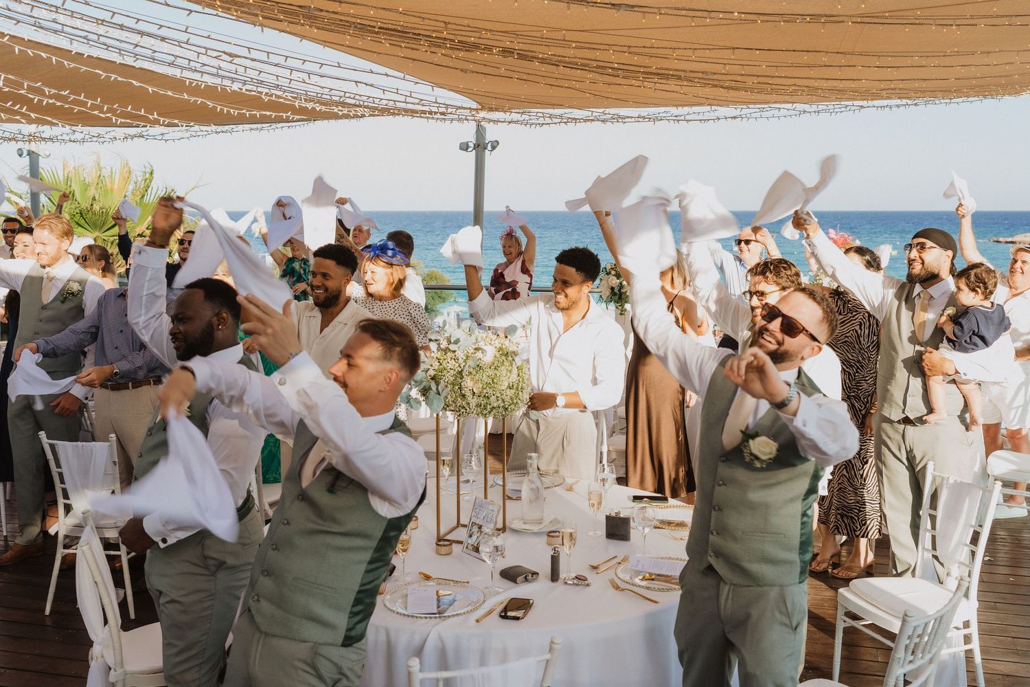 Wedding guests celebrate under a beachside tent, waving white napkins in the air during a festive reception.