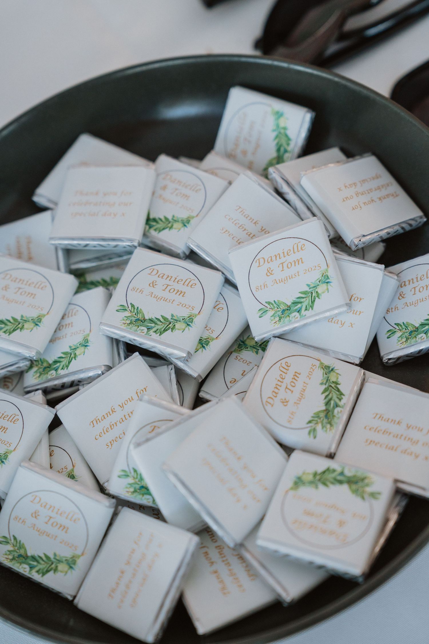 Wedding favor chocolates wrapped in white paper with green leaf designs arranged in a dark serving bowl.