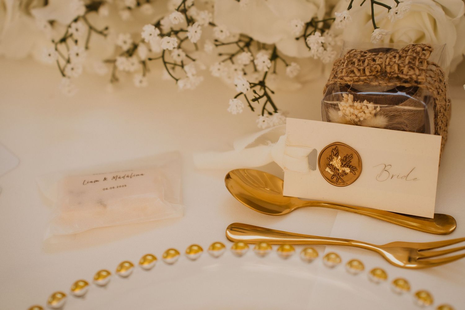 Elegant wedding place setting with gold cutlery, wax seal name card, and white floral centerpiece on white tablecloth.