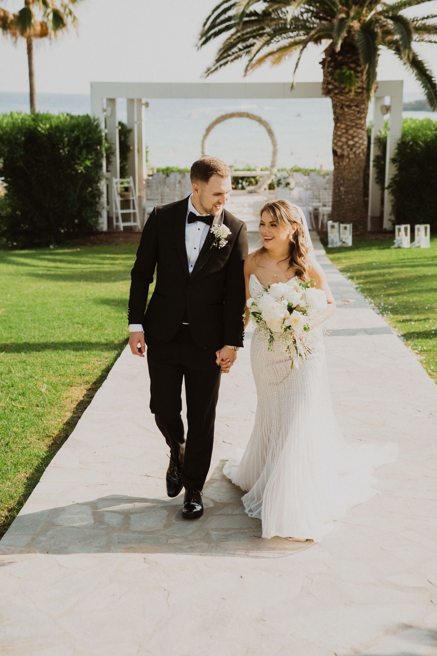 Bride and groom walk hand in hand along garden path with palm trees and arch backdrop.