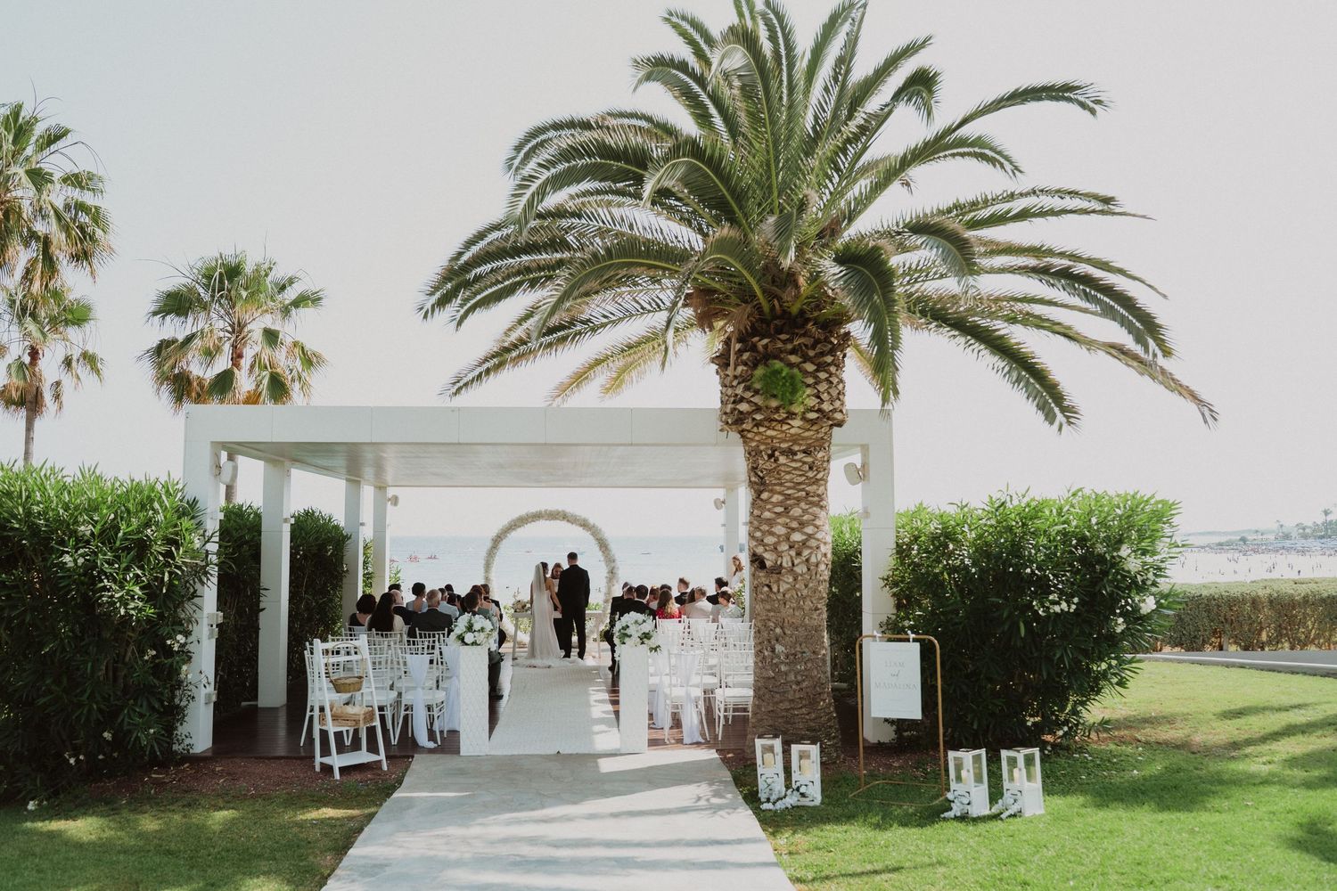 Outdoor wedding ceremony under white pavilion surrounded by palm trees and lush greenery.