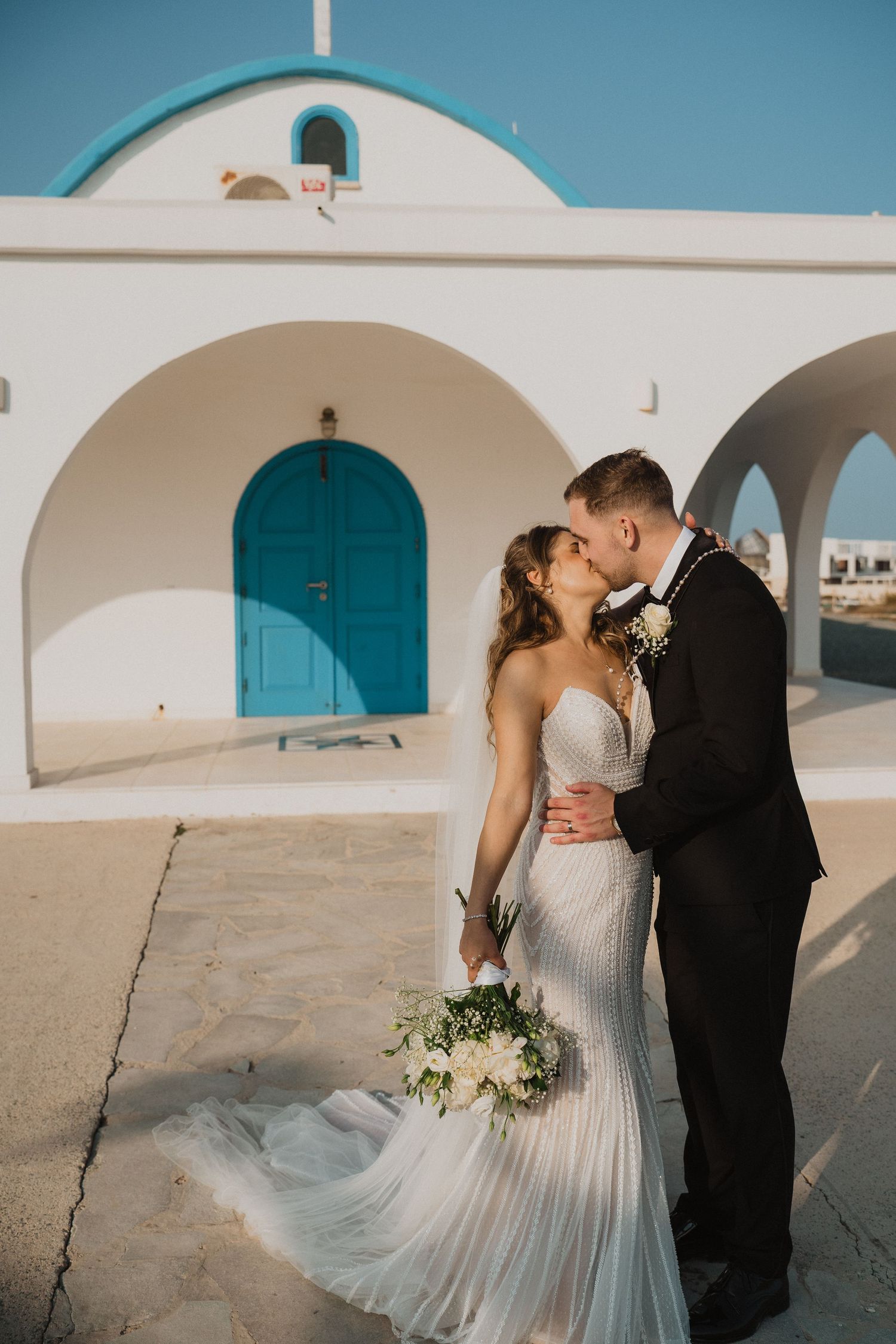 Bride and groom share a kiss outside a white Santorini chapel with a turquoise door on their wedding day.