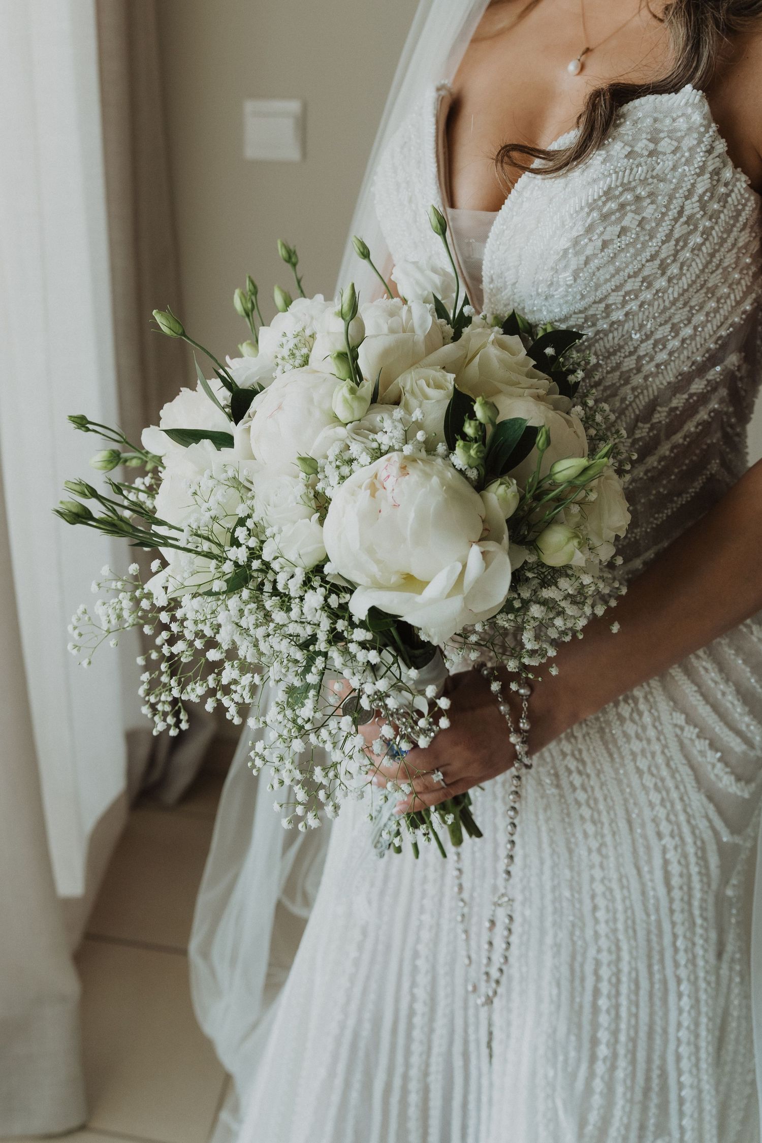 Bride holding a white wedding bouquet of peonies, roses, and baby's breath in a beaded gown.