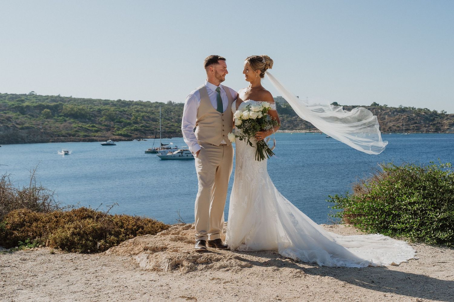 A couple in wedding attire stands together on a scenic coastal cliff overlooking blue ocean waters.