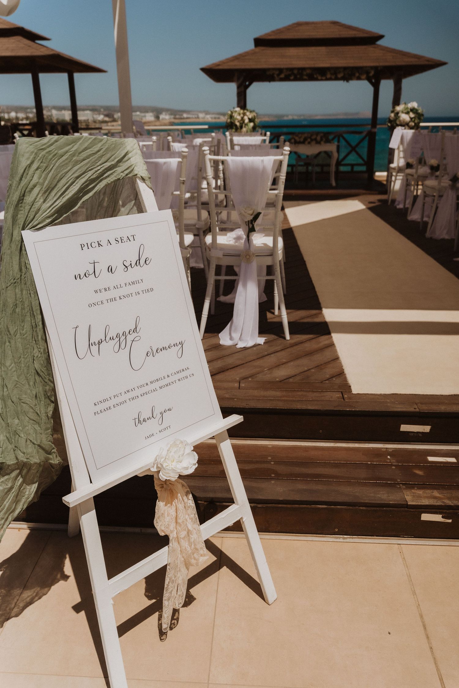 White wooden easel displays elegant wedding signage with beige bow accent at an outdoor beach venue with umbrellas.