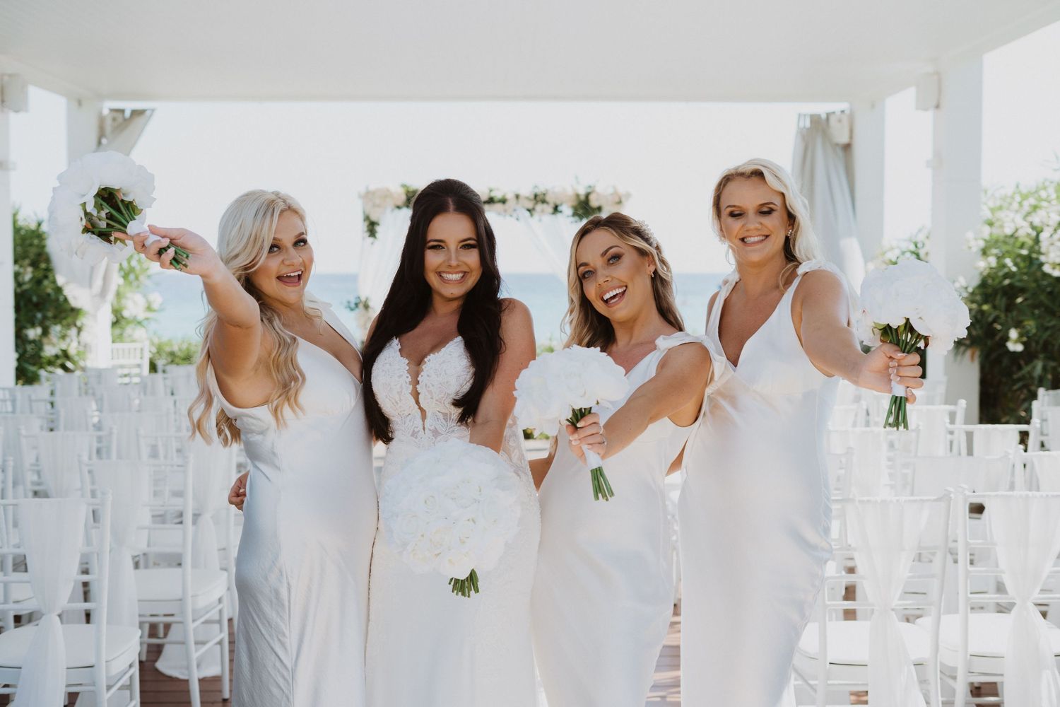 Bridesmaids in white dresses posing under an elegant white wedding ceremony structure.