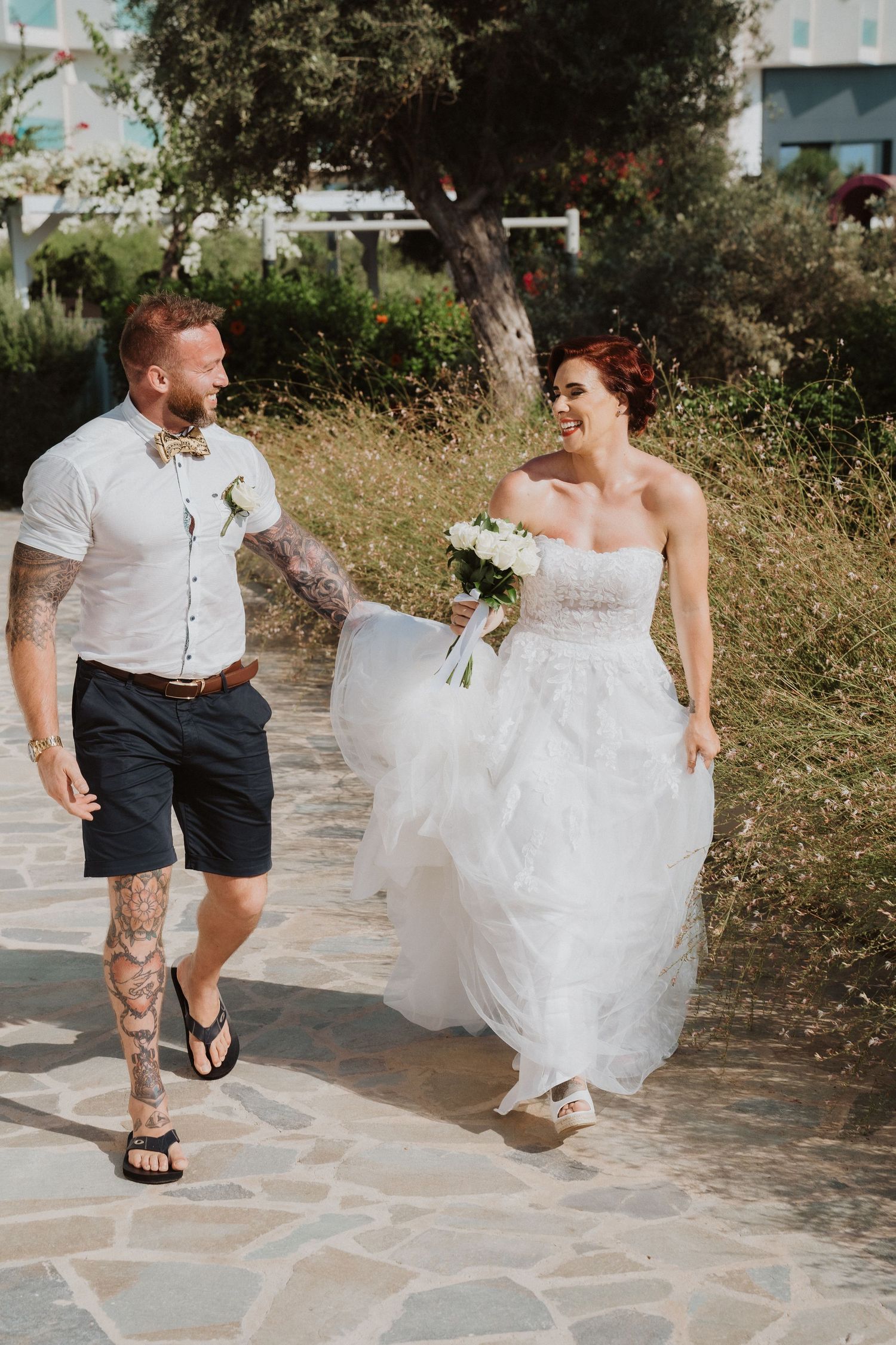 Newlyweds walk together on stone path, bride in flowing white dress.