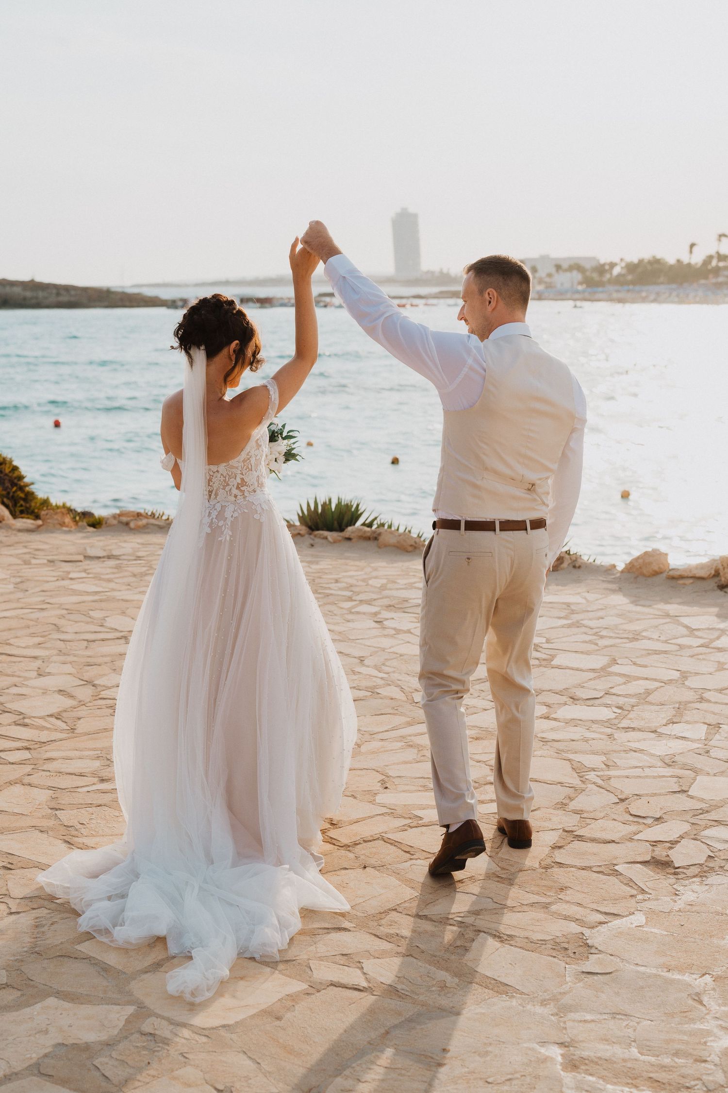 A couple in wedding attire dances together on a stone patio overlooking a scenic coastal beach at sunset.