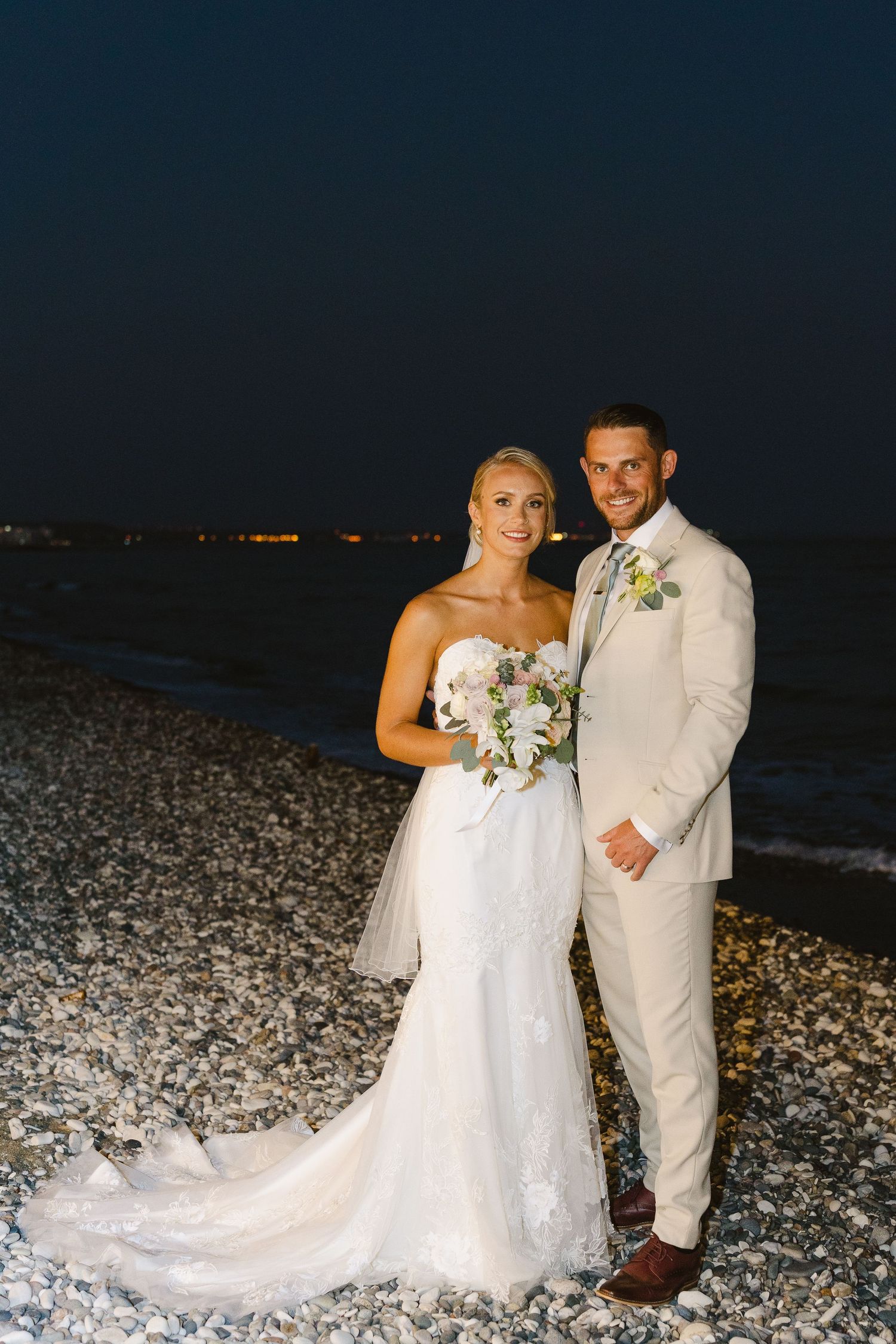 Newlyweds pose for a romantic beachside portrait at night in elegant wedding attire.