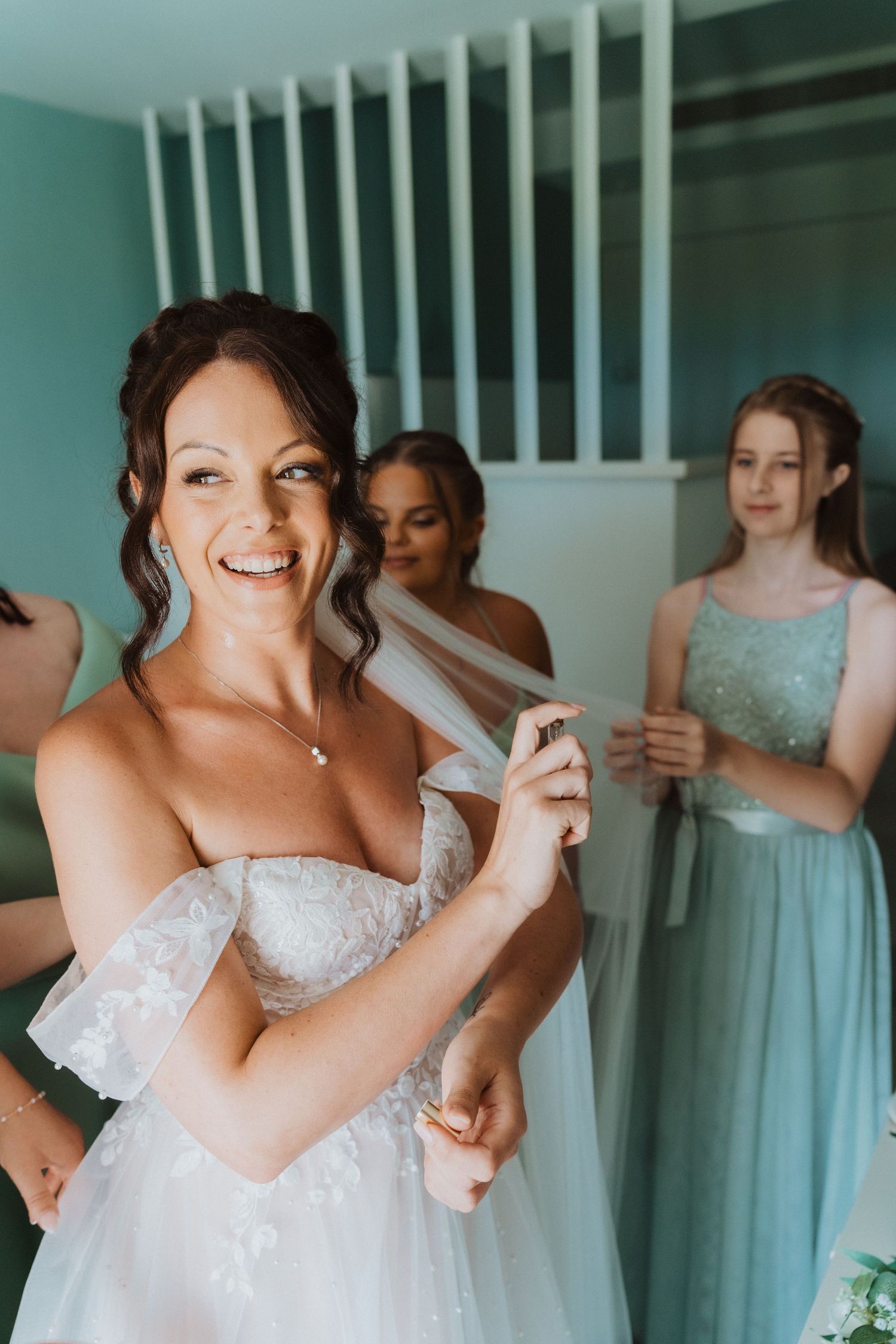 Joyful bride laughing while getting ready with bridesmaids in mint green dresses in bridal suite.