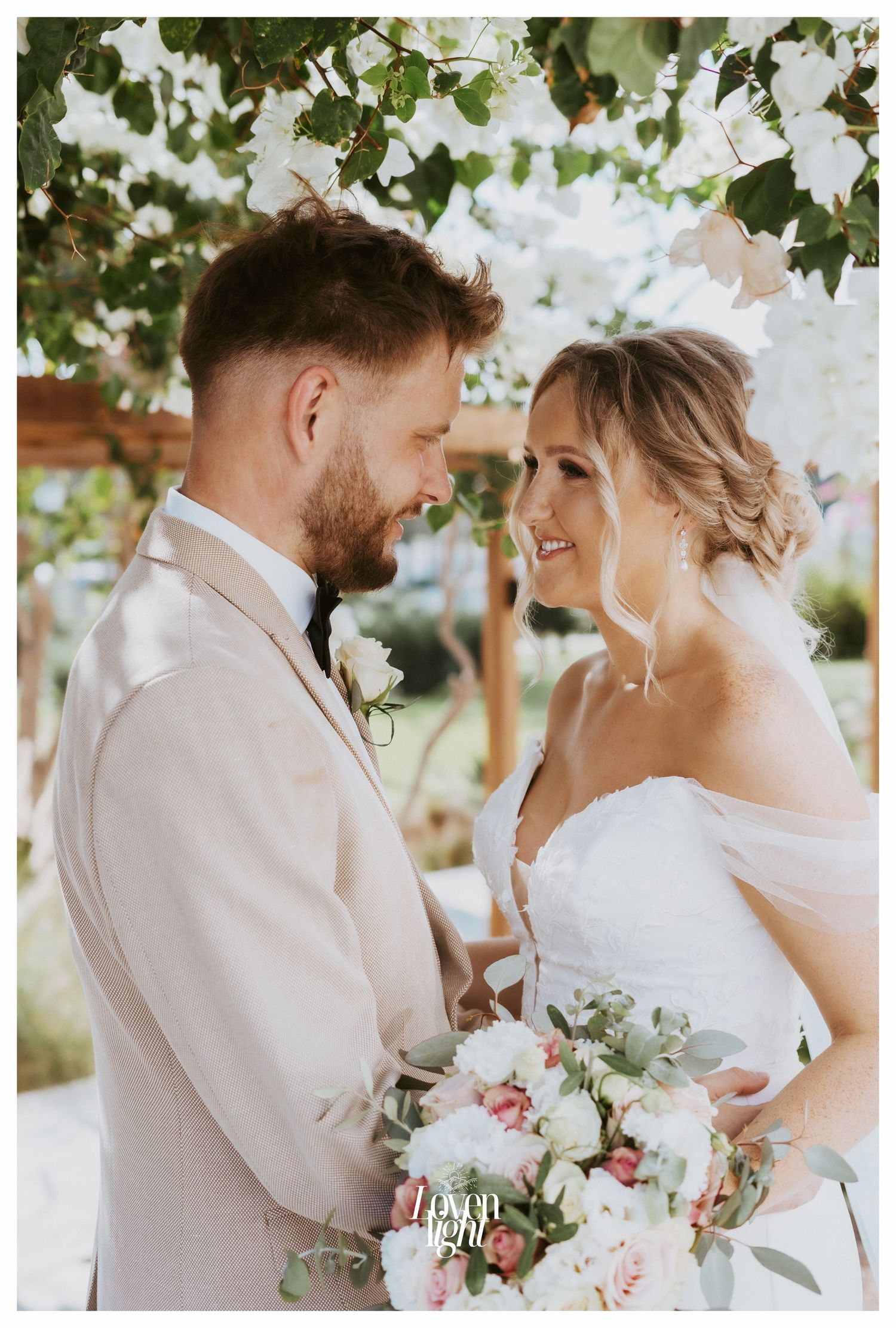 A romantic wedding moment captured under white flowers with the couple sharing a loving glance and holding a pink bouquet.