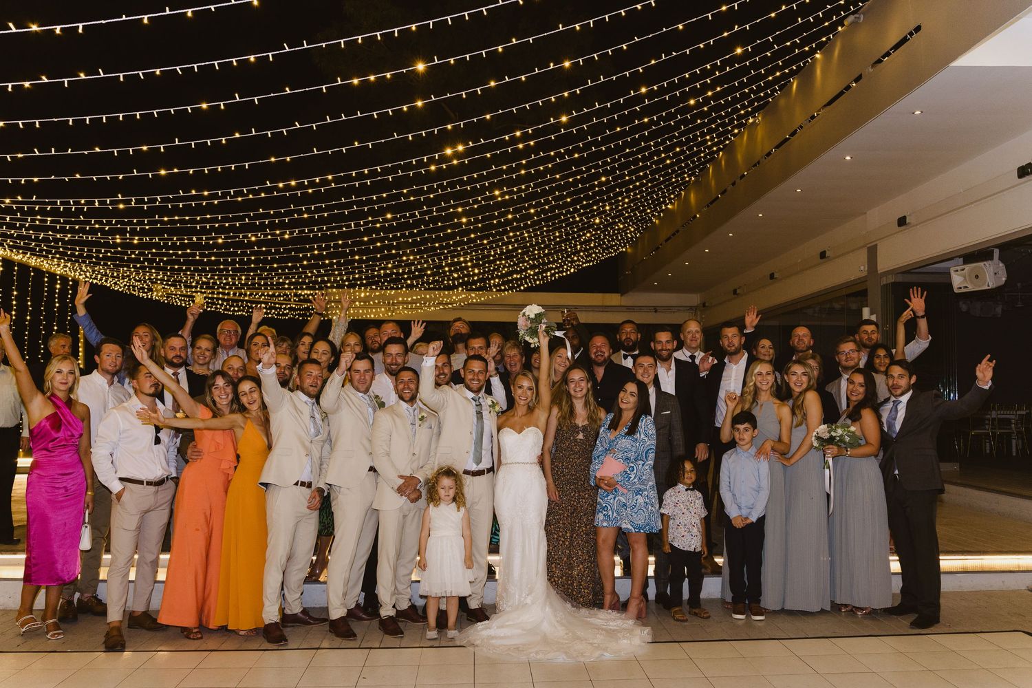 Large wedding group photo showing guests in colorful attire and groomsmen in beige suits under string lights.