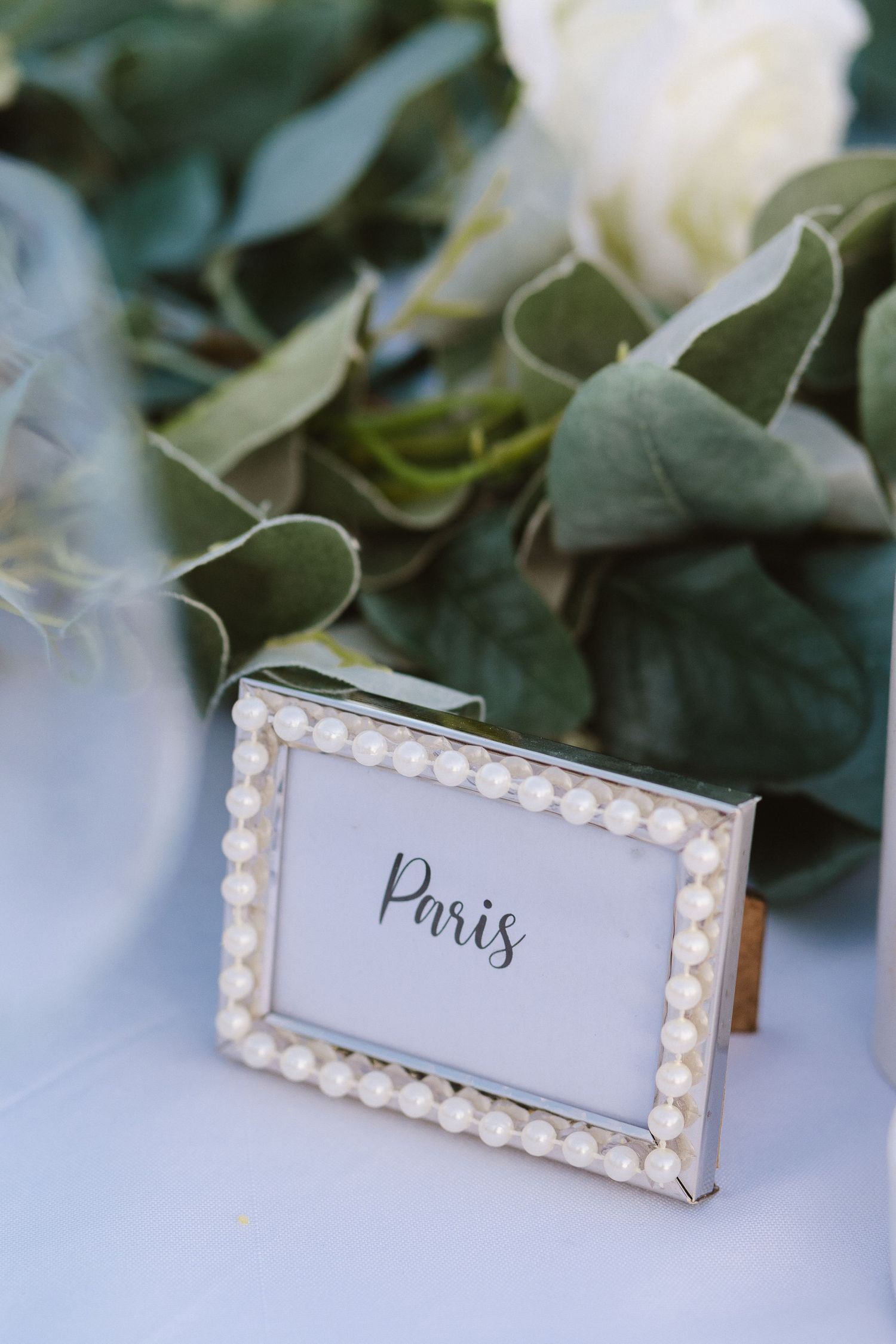 Pearl-bordered frame displaying 'Paris' text amid white roses and green foliage on an elegant table setting.
