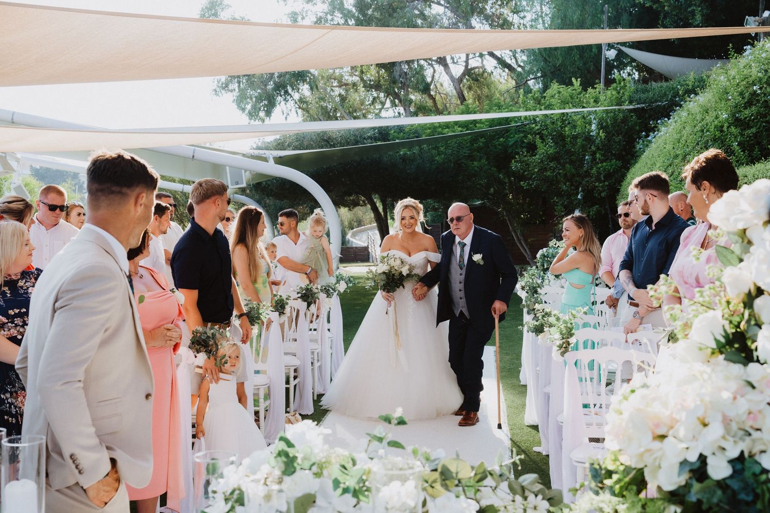Wedding guests gathered under a white canopy for an outdoor ceremony with elegant white flower arrangements.